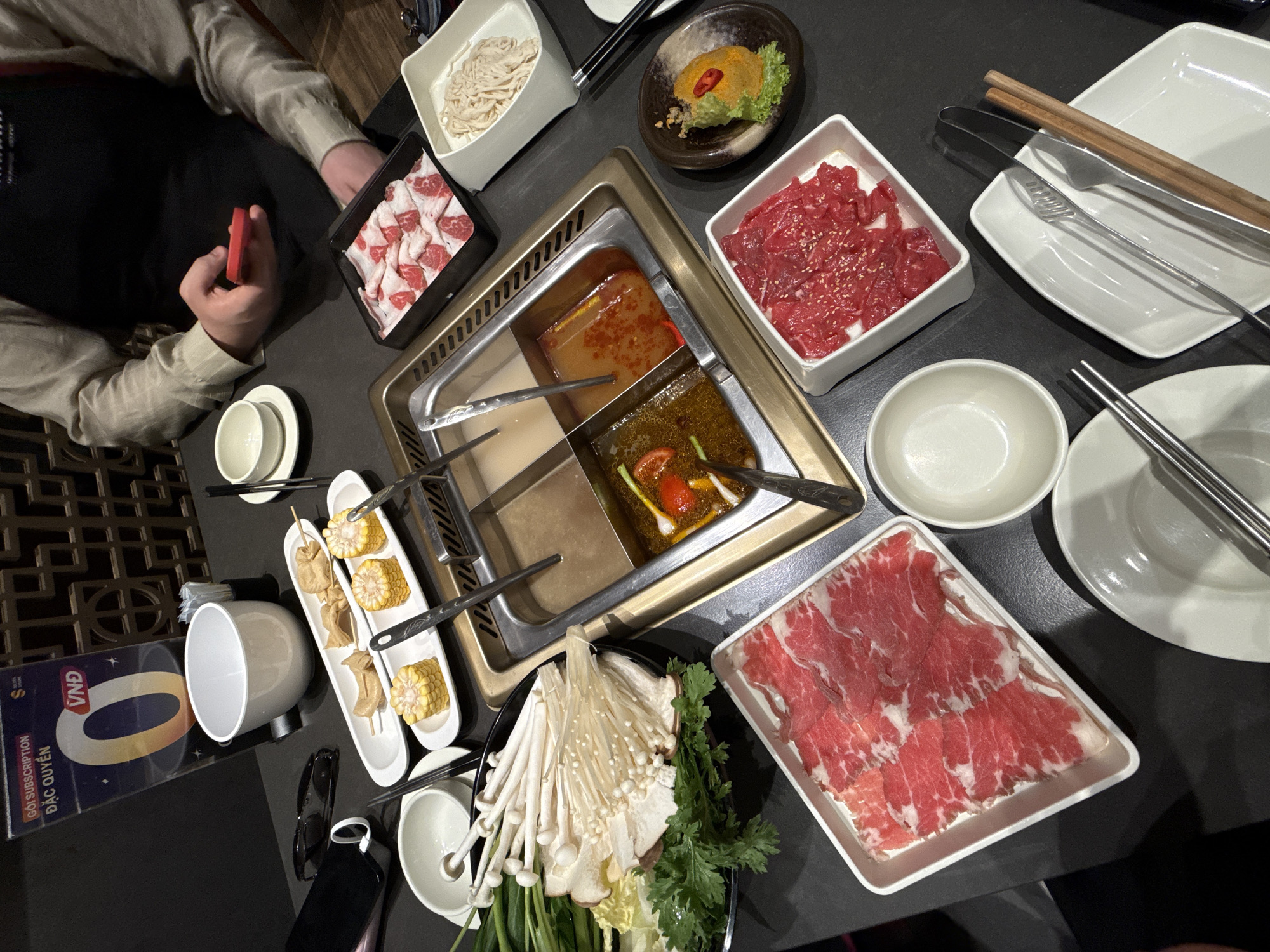 The media file presents a top-down view of a hot pot dining experience, likely within a restaurant setting in Nha Trang, Vietnam. A rectangular hot pot cooker with several broth compartments, featuring different broths (one with visible tomatoes and green onions), occupies the center of a dark table.
On the left, a person is partially visible, wearing a light-colored long-sleeved shirt and a black apron, holding a small red object in their hand. Around the hot pot, various white ceramic dishes are filled with ingredients ready for cooking. These include two plates of thinly sliced raw beef, one sprinkled with sesame seeds. Another plate holds an assortment of enoki mushrooms, other mushroom varieties, and fresh leafy greens such as napa cabbage and spring onions. There is also a dish containing thin white noodles or pasta, and a long plate with pieces of corn on the cob and skewered items resembling fish cakes. A small, dark bowl contains a yellow-orange paste, possibly a dipping sauce or minced shrimp, garnished with a red chili and green herb.
The table is set with white ceramic bowls and plates, metal and wooden chopsticks, and serving tongs. Personal items like sunglasses and a dark object, possibly a phone or power bank, are also present. In the background on the left, a decorative wooden lattice pattern adds to the ambiance. A promotional sign is visible with the text "VNA" and "GỌI SUBSCRIPTION ĐẶC QUYỀN," which translates to "EXCLUSIVE SUBSCRIPTION," confirming the Vietnamese location. The scene depicts a communal and enjoyable meal. The time of day is not explicitly indicated but appears to be a typical dining hour indoors.