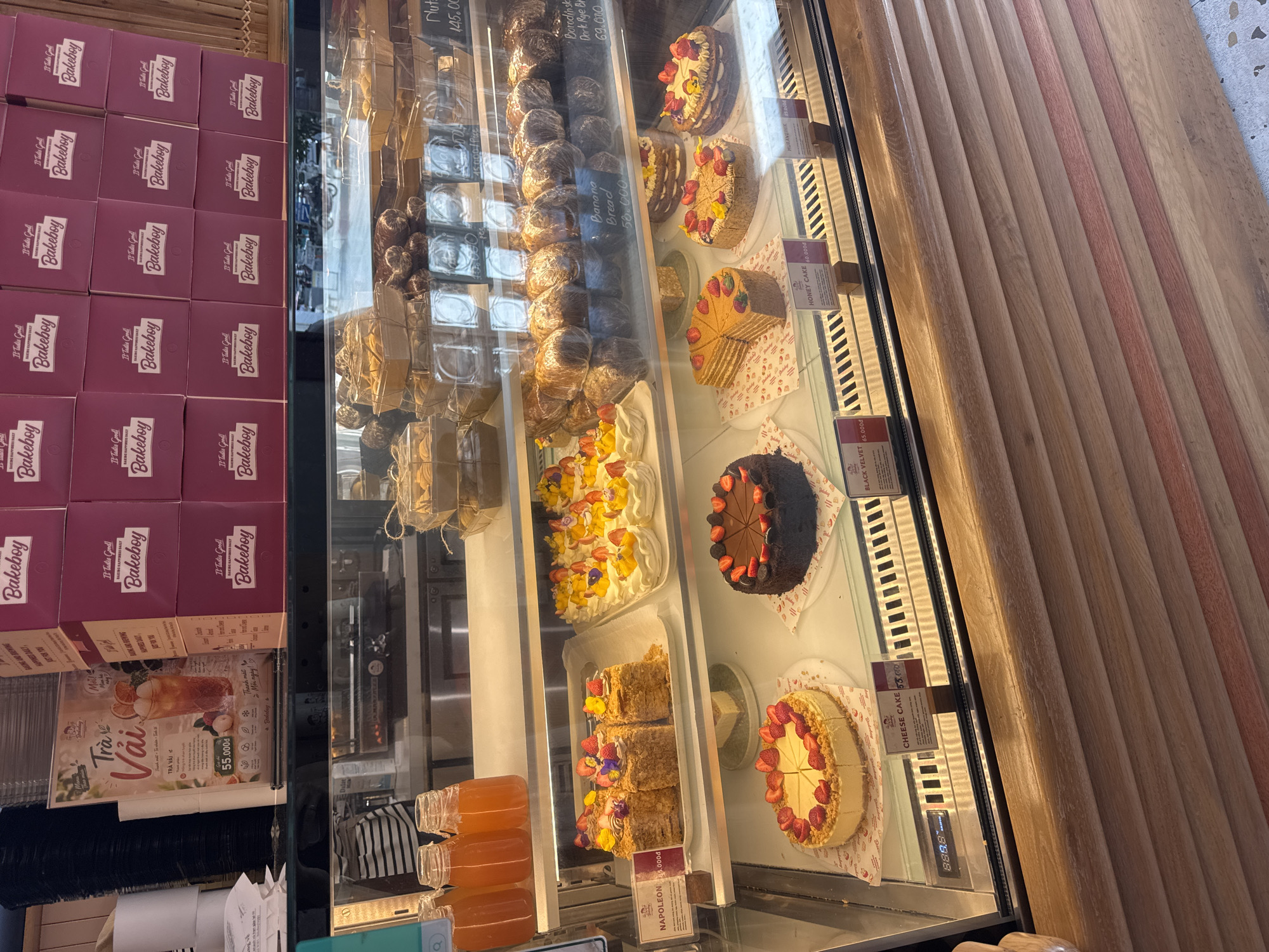 The image shows a display case in a bakery or cafe in Bac Nha Trang Ward, Vietnam. The display case is filled with various baked goods, including cakes, pastries, and bread. In the foreground, there are several cakes on display, including a strawberry cheesecake, a chocolate cake, and a honey cake. Behind these, there are more cakes and pastries, as well as loaves of bread wrapped in plastic. The display case is well-lit, and the baked goods appear fresh and appetizing.
The background of the image shows shelves filled with boxes, likely for take-away orders. There is also a sign with Vietnamese text advertising "Trà Vải" (lychee tea). The overall impression is of a clean, modern establishment selling a variety of sweet treats.
