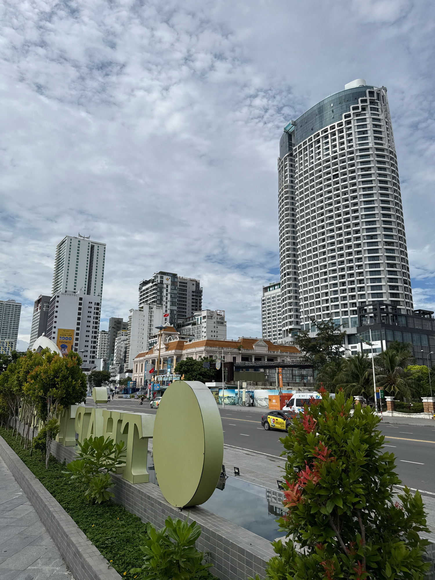 The image shows a street view in Nha Trang, Vietnam. In the foreground, a large, light green circular sign is visible, alongside a row of similarly colored large block letters spelling out a word, likely a business name or location identifier. These are set against a backdrop of lush green landscaping and a walkway. Beyond the landscaping, a street runs parallel to the walkway. On the street, there are several vehicles including a yellow taxi, a white van, and a motorcycle. In the background, a dense collection of modern, multi-story buildings dominate the skyline. One particularly tall, curved skyscraper with a white facade and dark tinted windows stands prominently on the right. To the left, a white hotel with a prominent "NOVOTEL" sign is visible, along with other varied architectural styles of buildings. The sky is cloudy, with patches of blue peeking through. The overall scene suggests a bustling urban environment in a coastal city.