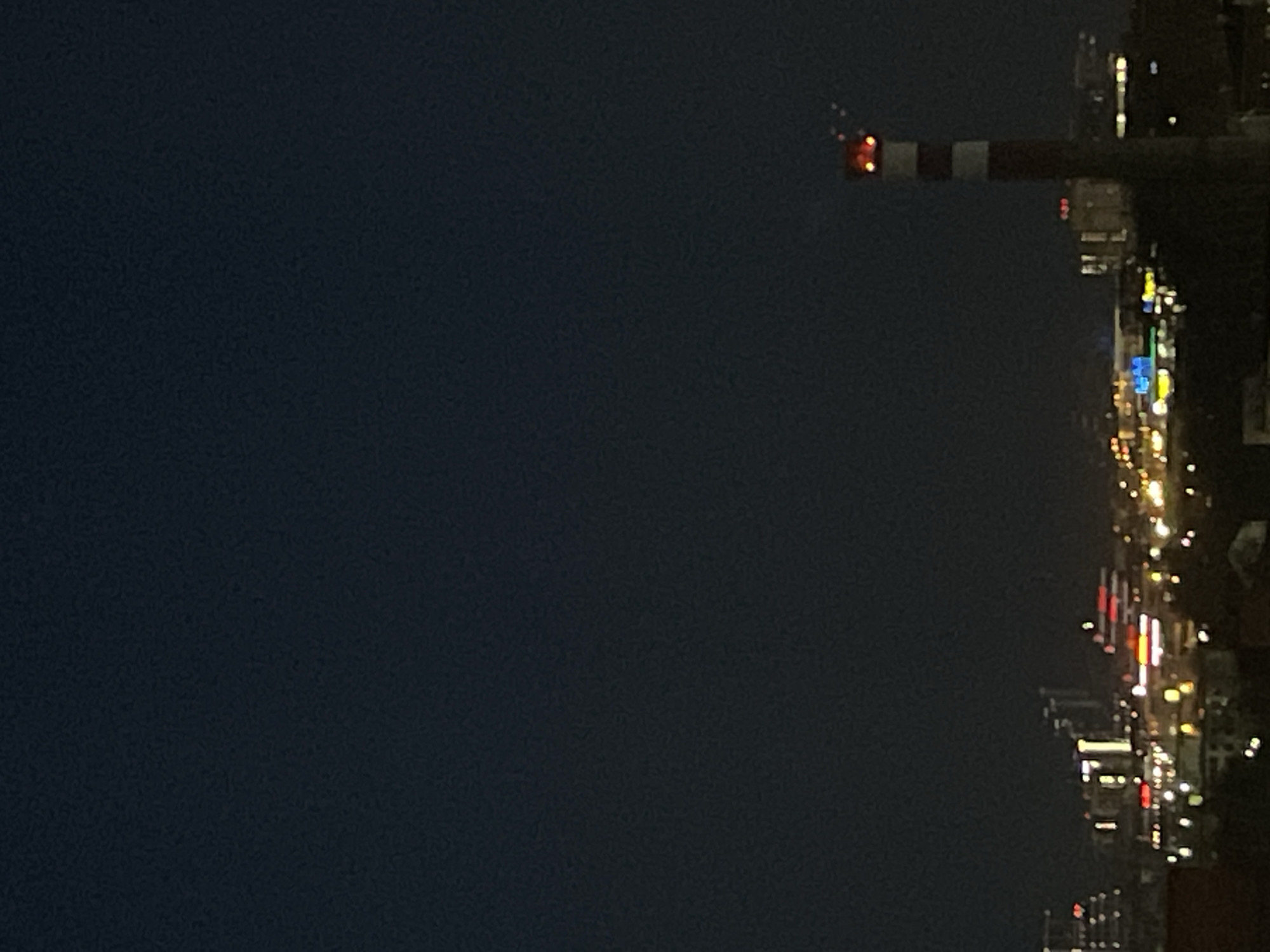 The image depicts a night view in what appears to be the city of Izhevsk, Russia. On the right side, city lights illuminate a street scene. A construction crane, marked with red and white stripes, is visible. The left side of the image is dominated by the dark night sky. The image is slightly blurred.