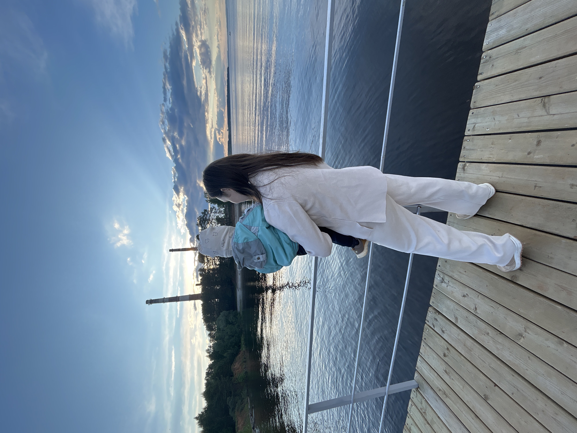 The image shows a woman in a white outfit holding a small child in a blue jacket and patterned hat at a pier in Chaikovsky, Russia. They are standing by a metal railing, with the water and landscape visible in the background. In the distance, two tall industrial smokestacks rise above the trees. The sky is partially cloudy with the sun shining through. The pier is made of wooden planks.