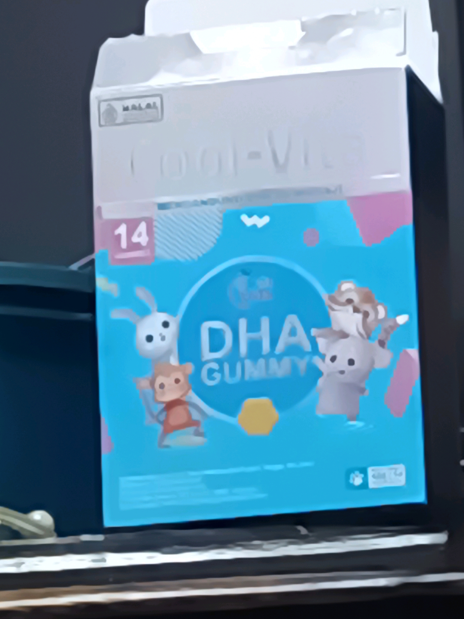 The image is a medium-shot photograph of a box of "DHA GUMMY" vitamins, likely taken indoors on a table. The box is predominantly teal and white, with cartoon animal illustrations of a rabbit, a monkey, an elephant, and a tiger on the front. The text "DHA GUMMY" is clearly visible in large, white letters. A small pink rectangle with the number "14" inside is positioned on the left side of the box. A "MALAL" logo is present in the upper left corner of the box. The box is slightly blurred, suggesting it might be out of focus or the image was taken quickly. The background is dark, with some hints of office equipment or furniture. The overall impression is that this is a product shot or a casual photograph of the product. There is no identifiable location in the city of South Jakarta, Indonesia, visible in the image.