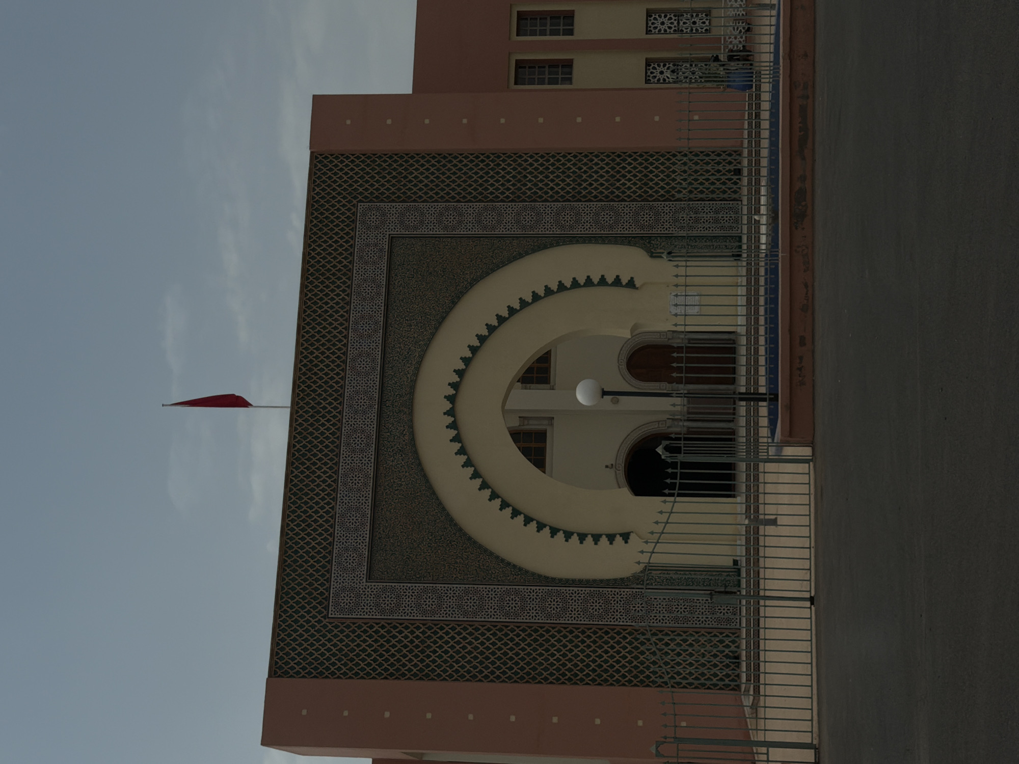 The image shows the exterior of a building with intricate, geometric mosaic tilework around a large archway. The building facade is painted in earthy tones, with accents of pink and patterned tile. A red flag flies from a pole atop the building. A metal fence runs across the foreground, separating the street from the building's entrance. The sky is a pale blue with some wispy clouds. The scene appears to be located in Marrakesh, Morocco, likely a public building or a significant historical site given the architectural style.