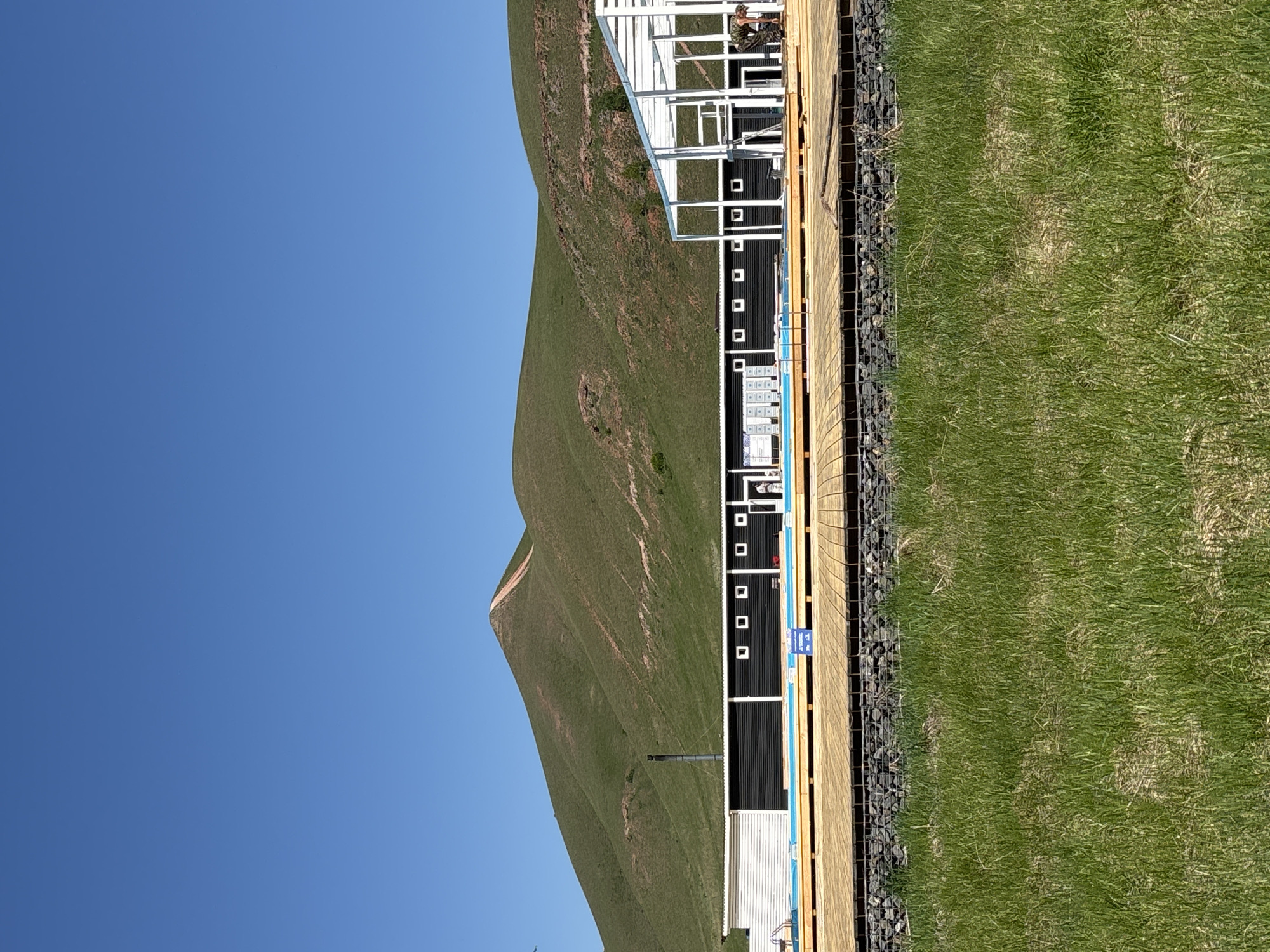 The image, correctly oriented, displays a long, low-profile building or series of modular units situated at the base of a large, verdant hillside under a clear, bright blue sky.
The main subjects are the building, the hill, and a person. The building appears modular, featuring predominantly black sections with numerous small, square white windows or panels. One end of the structure includes a white, skeletal frame, possibly indicating ongoing construction or a covered area. A wooden platform or deck runs along the front of the building, and adjacent to it is a gabion wall, constructed from wire cages filled with rocks. In the foreground, a strip of green grass is visible.
A single person is present on the wooden platform, near the white framed section of the building. They are dressed in camouflage-patterned clothing and appear to be engaged in an activity, possibly working or inspecting the structure. A dark, vertical pipe-like object, resembling a chimney or vent, extends from one of the black modules.
The activity taking place seems to be related to construction, maintenance, or operation of the facility. The hillside is covered in green grass with patches of drier vegetation and a faint dirt path winding up its side.
Notable details include the bright, clear blue sky and strong sunlight, indicating a sunny day, likely morning or afternoon. The setting is outdoors, possibly in a rural or semi-rural area near Дрокино, Russia, given the natural landscape. A small blue sign with white text is visible on the wooden platform, displaying the Cyrillic word "ВЫХОД" (meaning "EXIT").