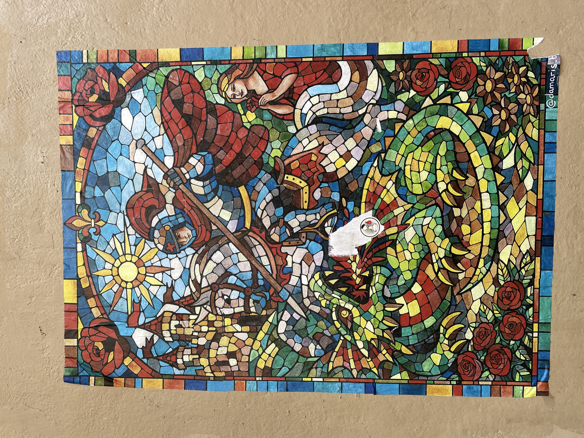 A vibrant, mosaic-style artwork, resembling stained glass, is affixed to a textured, light brown wall in Barcelona, Spain. The artwork depicts a fantastical scene, possibly illustrating the legend of Saint George and the Dragon.
On the left side, a figure reminiscent of a knight or saint is shown in a dynamic pose, dressed in red and blue segmented armor. This figure holds a long weapon, possibly a spear or sword, pointed towards a large creature. Above this figure, a radiant sun motif with pointed rays is prominent, alongside decorative red roses and a blue fleur-de-lis.
In the upper-middle section, a reclining or sleeping female figure, perhaps a maiden or princess, is depicted with flowing red garments and blonde hair, her form composed of segmented shapes characteristic of the stained-glass aesthetic.
Dominating the right side of the composition is a large, green, scaly creature, strongly resembling a dragon. Its body is intricately patterned with shades of green, yellow, and red, and it features sharp teeth and clawed limbs. This creature appears to be engaged in conflict with the knightly figure. The top-right corner also features several red roses and brown floral patterns amidst green leaves.
The entire artwork is framed by a colorful mosaic border. The piece itself, made of paper or fabric, shows minor creases and wear. A small white sticker with a red heart icon partially covers a portion of the dragon's body. In the top right corner, vertically aligned, the text "@damaris_b" is visible, likely an artist's signature or social media handle. The textured wall provides a neutral backdrop to the colorful and intricate street art.