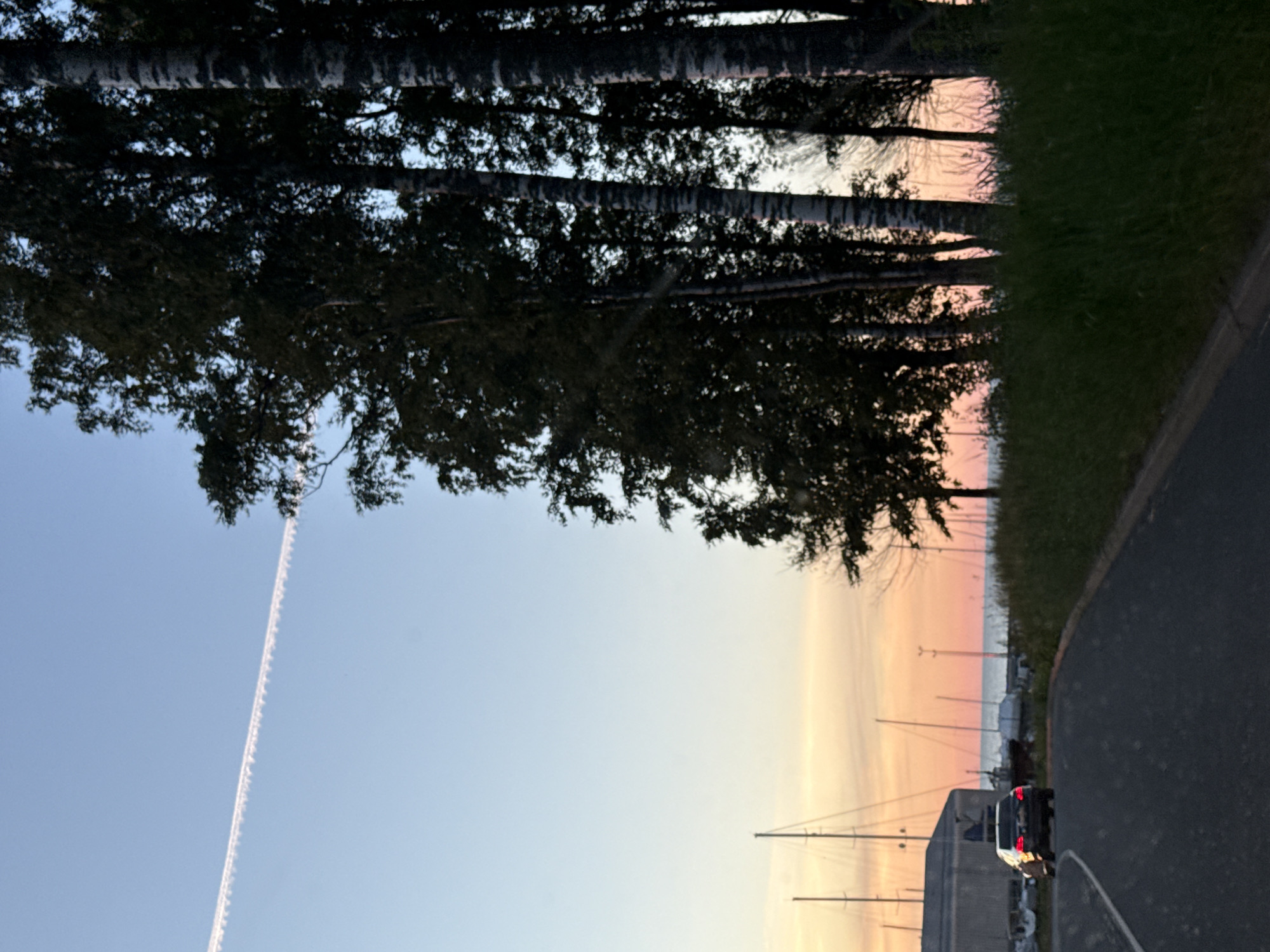 A car drives down a road next to a grassy embankment and a line of birch trees. In the background, sailboats are moored in a harbor, with a building behind them. The sky is clear blue with a faint contrail from an airplane. The sun is setting, casting a warm glow on the horizon. This scene appears to be from the Pirita district of Tallinn, Estonia, a popular area for recreational activities and a marina.