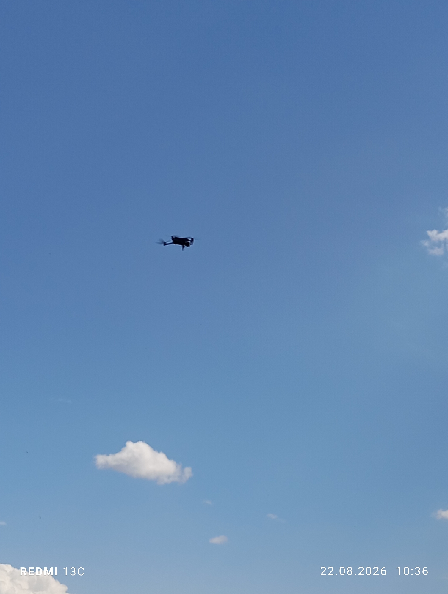 A black drone is captured in mid-flight against a clear blue sky. The drone is positioned in the upper-middle portion of the frame, appearing as a small dark silhouette against the expansive sky. A single, small white cloud is visible in the lower-middle section of the image, to the left of the drone. The bottom left corner of the image displays the text "REDMI 13C" and the bottom right corner shows "22.08.2026 10:36". The scene is devoid of any people or discernible landmarks suggesting the Amirsay ski resort in Uzbekistan.