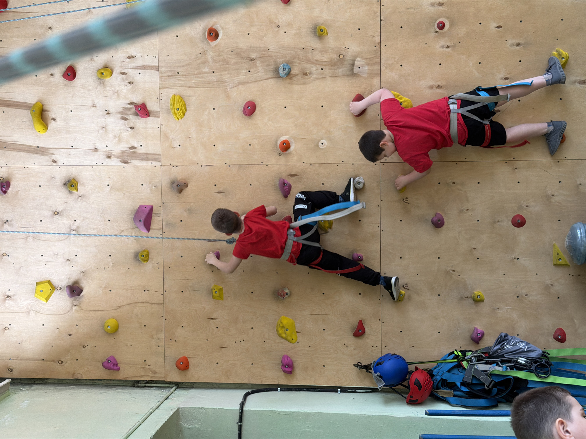 Two boys in red t-shirts and climbing harnesses are climbing on an indoor climbing wall. The wall is made of plywood and has various colored climbing holds attached to it. The boy on the left is wearing black pants and black sneakers. The boy on the right is wearing black shorts and gray sneakers. Climbing equipment is scattered on the floor next to the wall, including harnesses and helmets. Part of a man's head is visible in the lower right corner. A rope extends from the top left of the image to the wall. The location is Izhevsk, Russia.