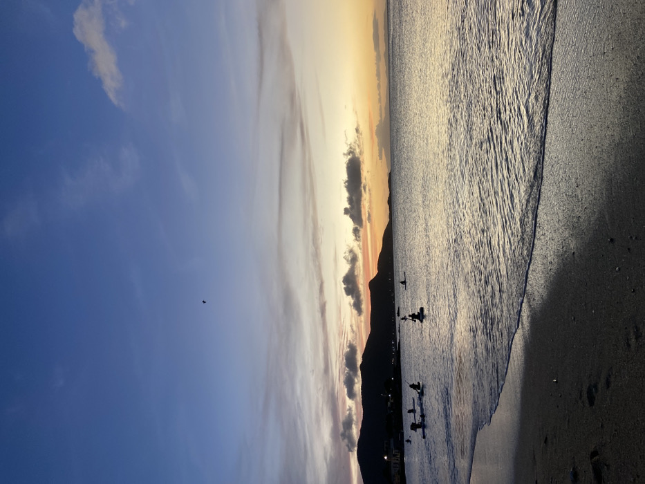 A scenic view features a beach at sunset. The sky displays a gradient of blues and oranges with wispy clouds. The sea reflects the sunset's colors, rippling gently towards the dark sandy beach. Silhouetted figures stand or float in the water near the shoreline. A dark mountain range forms a backdrop along the horizon. A tiny bird flies high in the clear blue sky.