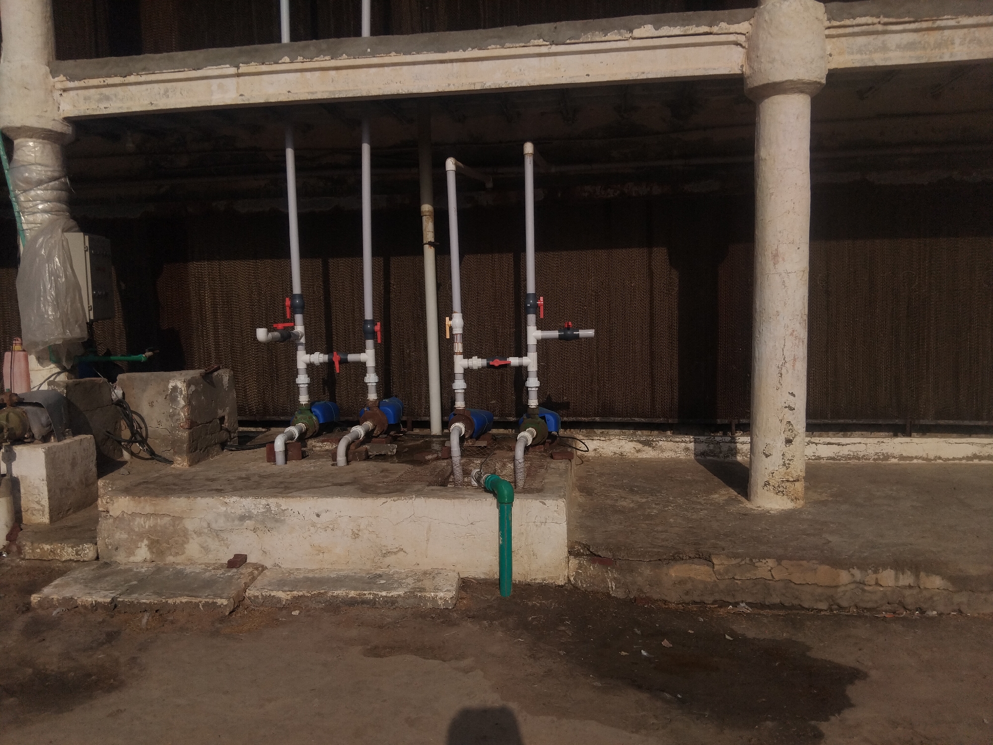 This is a medium shot, eye-level, outdoor photograph taken during the day in Haveli Jhangranwali, Pakistan. The scene depicts the operational infrastructure of a cooling system, likely for a poultry farm or similar agricultural facility. In the foreground, there is a concrete ground surface with some dark, wet patches and debris. Behind this, a raised concrete platform, weathered and cracked, supports various components of the cooling system.
The most prominent elements are a series of pipes and valves. White PVC pipes extend vertically from beneath the platform, branching out horizontally and fitted with red manual control valves. Attached to these are blue components, possibly pumps or water distribution units, with metal and PVC piping connecting them. A distinctive bright green PVC pipe is visible emerging from the platform and bending downwards.
The background is dominated by a large, dark, honeycomb-like structure, which appears to be a cooling pad or media, extending horizontally behind the piping. The structure is supported by an overhead concrete beam and a series of thick, cylindrical concrete columns, one of which is clearly visible on the right side of the image, showing signs of wear and tear. Another large concrete pipe, wrapped in white plastic sheeting, is visible on the left side of the image. The overall impression is of a functional, industrial outdoor space.