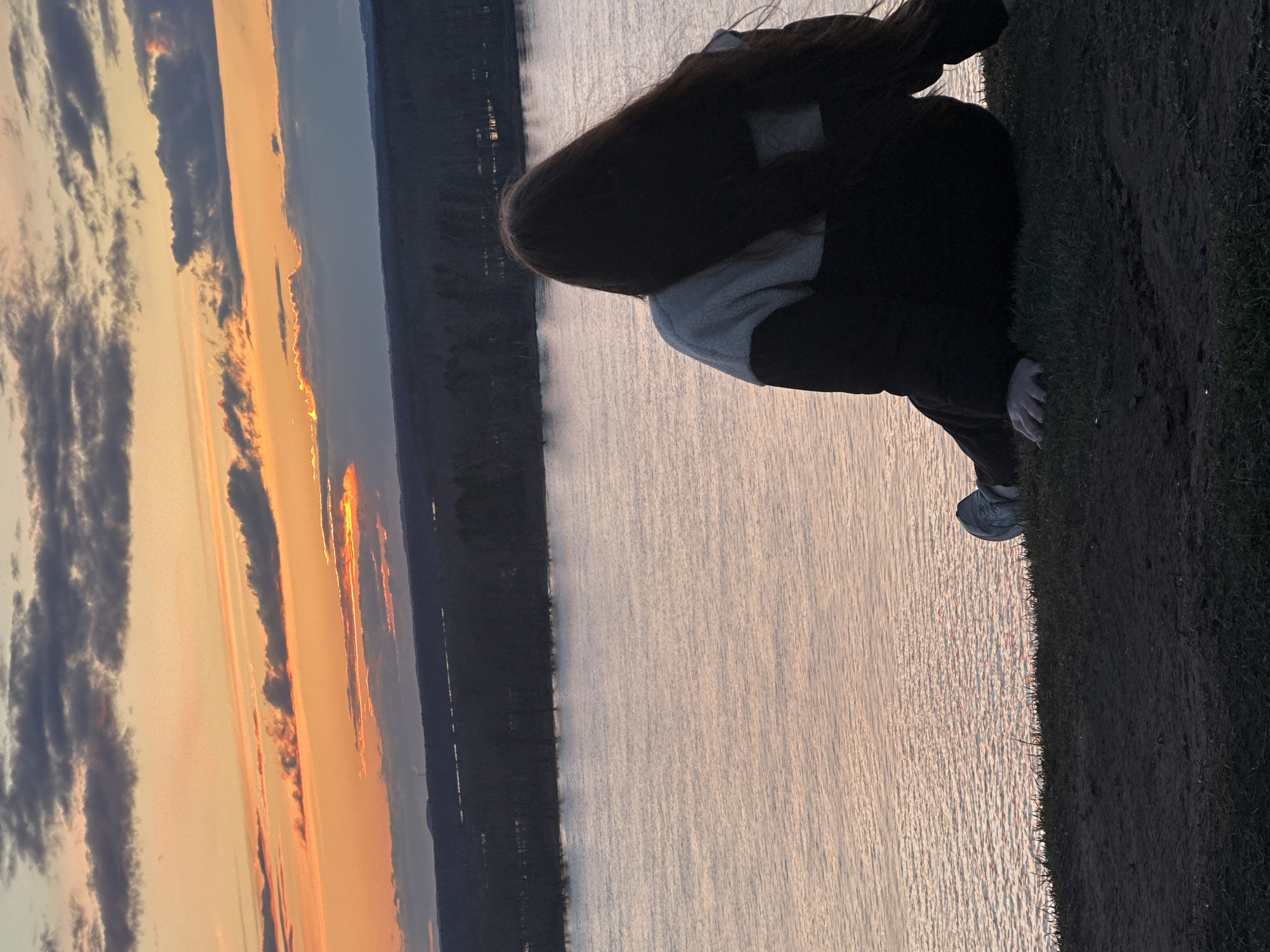 The image depicts a serene outdoor scene at sunset, focusing on a person observing a vast body of water. The subject is seen from behind, sitting or crouching on a grassy bank in the foreground. They have long, dark hair and are wearing a light-colored top under a dark outer layer, with dark pants and a grey or blue sneaker visible near the edge of the grass.
The sky is a prominent feature, displaying a vivid sunset with shades of orange, yellow, and red near the horizon, transitioning into cooler blues and grays above. Scattered dark clouds are illuminated by the setting sun, adding texture to the sky. The expansive body of water, possibly a river or a large lake, reflects the soft light of the sunset, creating gentle ripples and glimmers on its surface. Across the water, a dark, distant shoreline, likely composed of trees or landforms, stretches across the mid-ground. The immediate foreground consists of a dark green grassy area where the individual is positioned.
The activity suggests a quiet moment of contemplation or appreciation for nature's beauty as the day ends. The time of day is clearly sunset, marked by the rich colors of the sky and the soft light over the water. No visible text is present in the image. The setting suggests a natural landscape, potentially near a large water body in Ольховка, Russia.