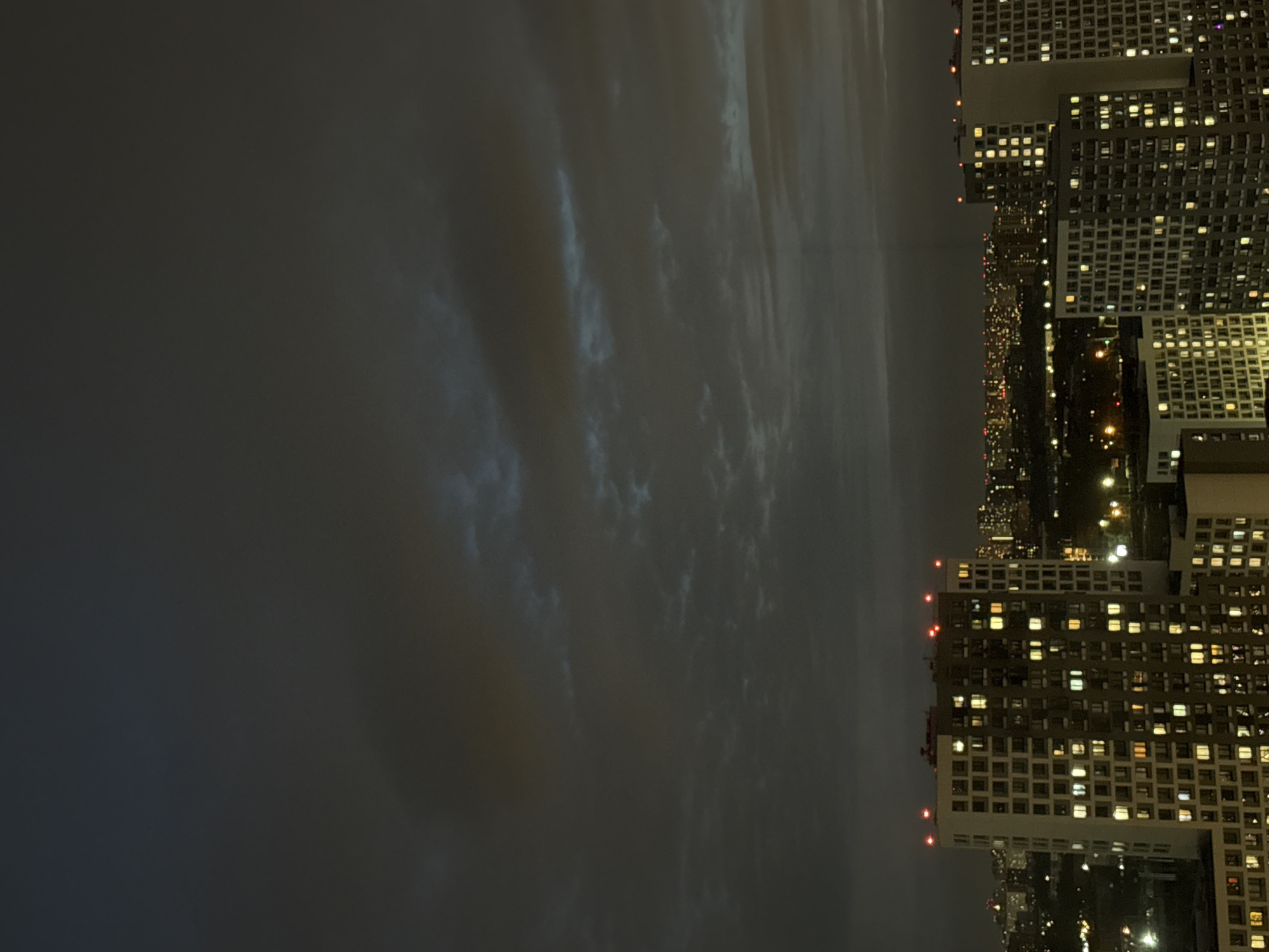 The image shows a cityscape at night. The right side of the image features tall buildings with numerous illuminated windows, suggesting residential or commercial spaces. Between the buildings, there are indications of streets with some light sources visible, possibly from streetlights or vehicle headlights. The left side of the image is dominated by a dark, possibly cloudy, night sky. The specific location within Lyubertsy, Russia, cannot be determined from the image alone. There are no people visible.