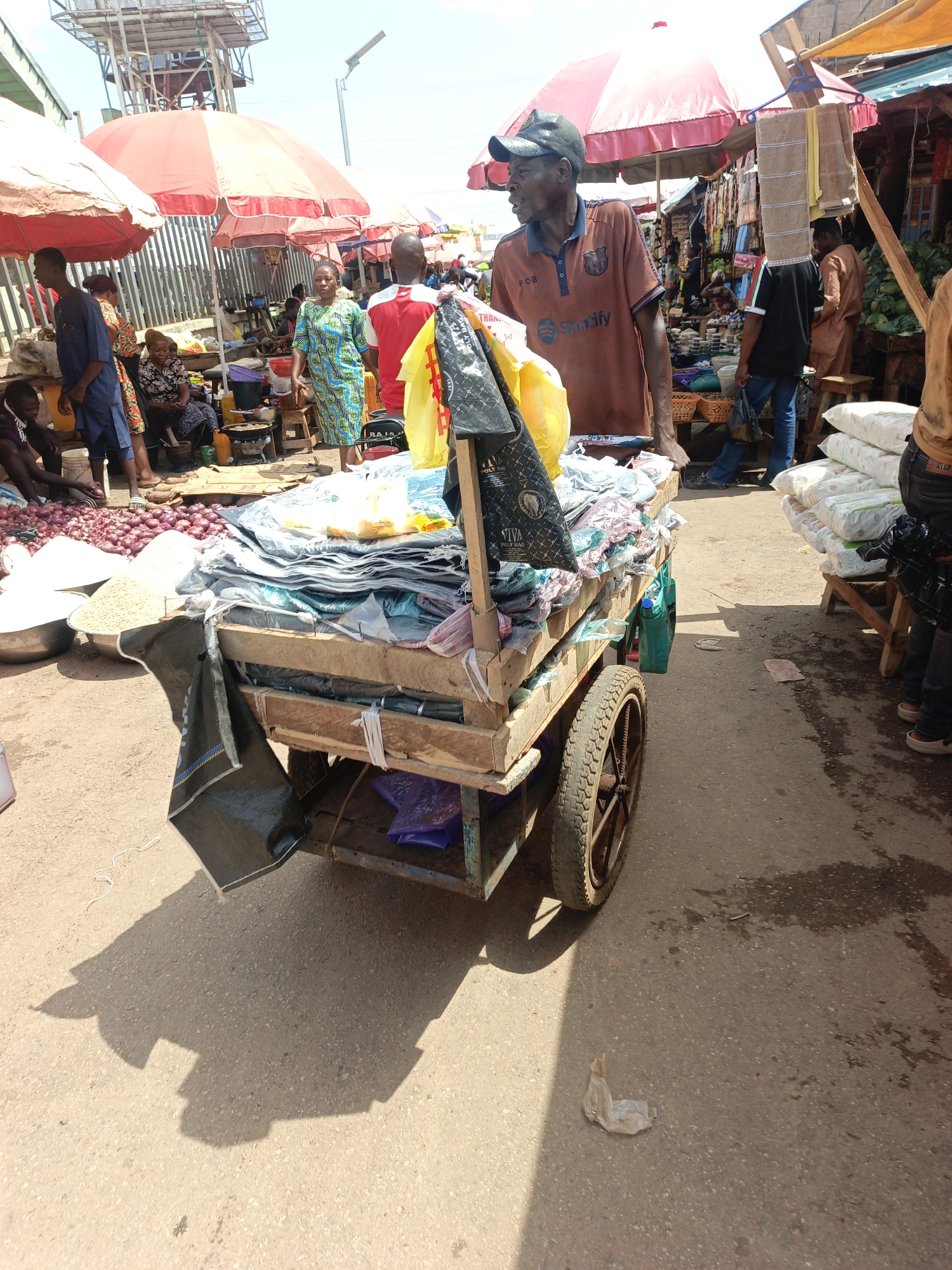 The image captures a vibrant street scene in Akwanga, Nigeria. The focal point is a middle-aged man wearing a brown polo shirt with a "Spotify" logo and a baseball cap, standing behind a heavily laden wooden cart. The cart is filled with various items wrapped in plastic, possibly goods for sale. The cart itself has two large wheels.
The man is looking to the right, seemingly engaging with someone or something out of frame. The background is bustling with activity, featuring several people, market stalls shaded by red and white umbrellas, and piles of goods like onions and bags of grain or rice. The environment suggests a busy marketplace, with a mix of vendors and customers. The ground is asphalt, showing wear and tear. The scene is bathed in bright sunlight, casting shadows from the cart and surrounding objects.