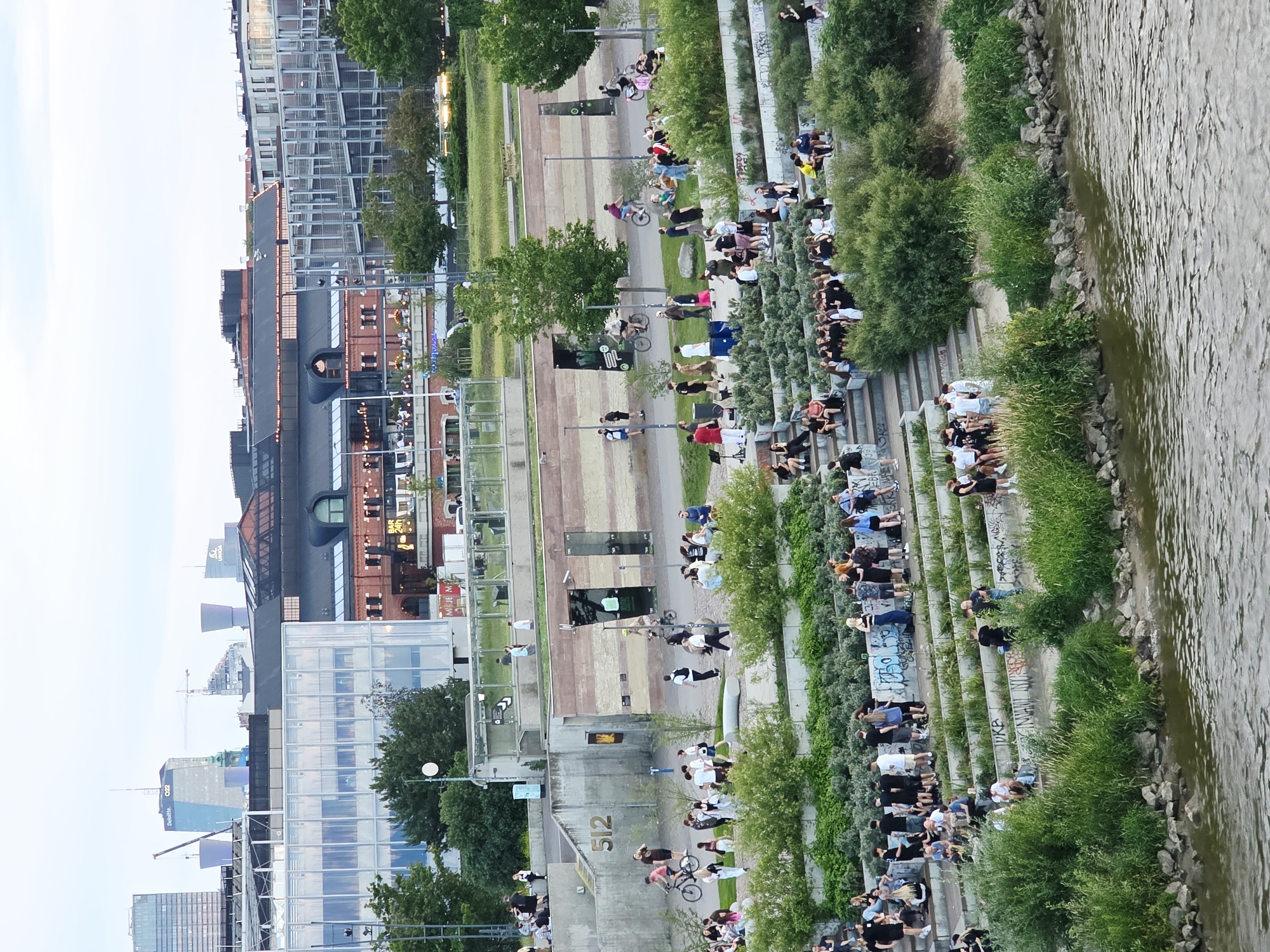 The image depicts an outdoor scene in Warsaw, Poland, likely on a warm day, with a focus on a tiered, landscaped area adjacent to a river. The architecture visible in the background suggests an urban setting with modern and older buildings, including a large structure with a red brick facade and a modern glass-fronted building.
In the foreground, a river occupies the bottom portion of the frame. The middle ground features a wide, multi-levelled embankment with grassy areas, concrete steps, and dense green foliage. Numerous people are scattered throughout this area, engaged in various activities. Some are sitting on the steps and grass, socializing in groups, while others are standing and walking. Several individuals are seen riding bicycles on a paved path that runs along the embankment. One person is on a scooter, and another appears to be playing an instrument in front of a small crowd. The overall atmosphere suggests a public space being utilized for recreation and leisure.
The upper part of the image shows a panorama of the Warsaw skyline, with several prominent buildings and structures, including skyscrapers and cranes, indicating active urban development. The sky is overcast, suggesting diffused lighting. The scene is captured from an elevated perspective, looking down towards the river and the embankment.