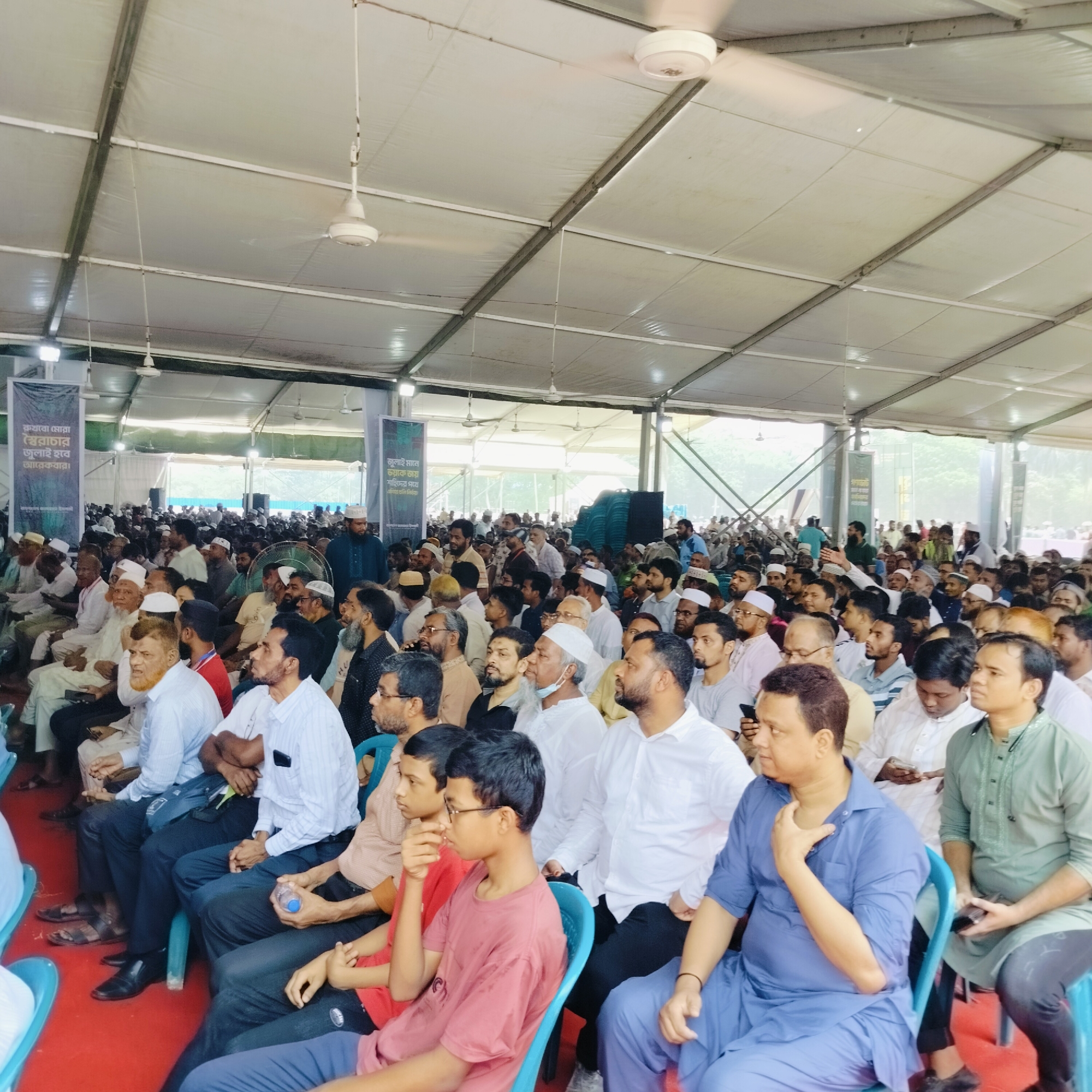 The image depicts a large gathering of people, predominantly men, seated in rows of blue plastic chairs within a tent-like structure. The environment suggests an event or conference setting, possibly religious or cultural in nature, considering the attire of many attendees, some of whom are wearing traditional Islamic clothing like taqiyahs (skullcaps). The red carpet on the floor adds a touch of formality to the space.
The individuals are of South Asian descent. They are diverse in age, from young boys to elderly men. Many are attentive, with their gazes directed towards a point beyond the frame. The tent overhead has ceiling fans, indicating an attempt to provide ventilation. Banners with text written in Bengali are visible in the background. Based on the presence of Bengali script and the overall appearance of the subjects, it is most likely that this image was taken in the city of Dhaka, Bangladesh.