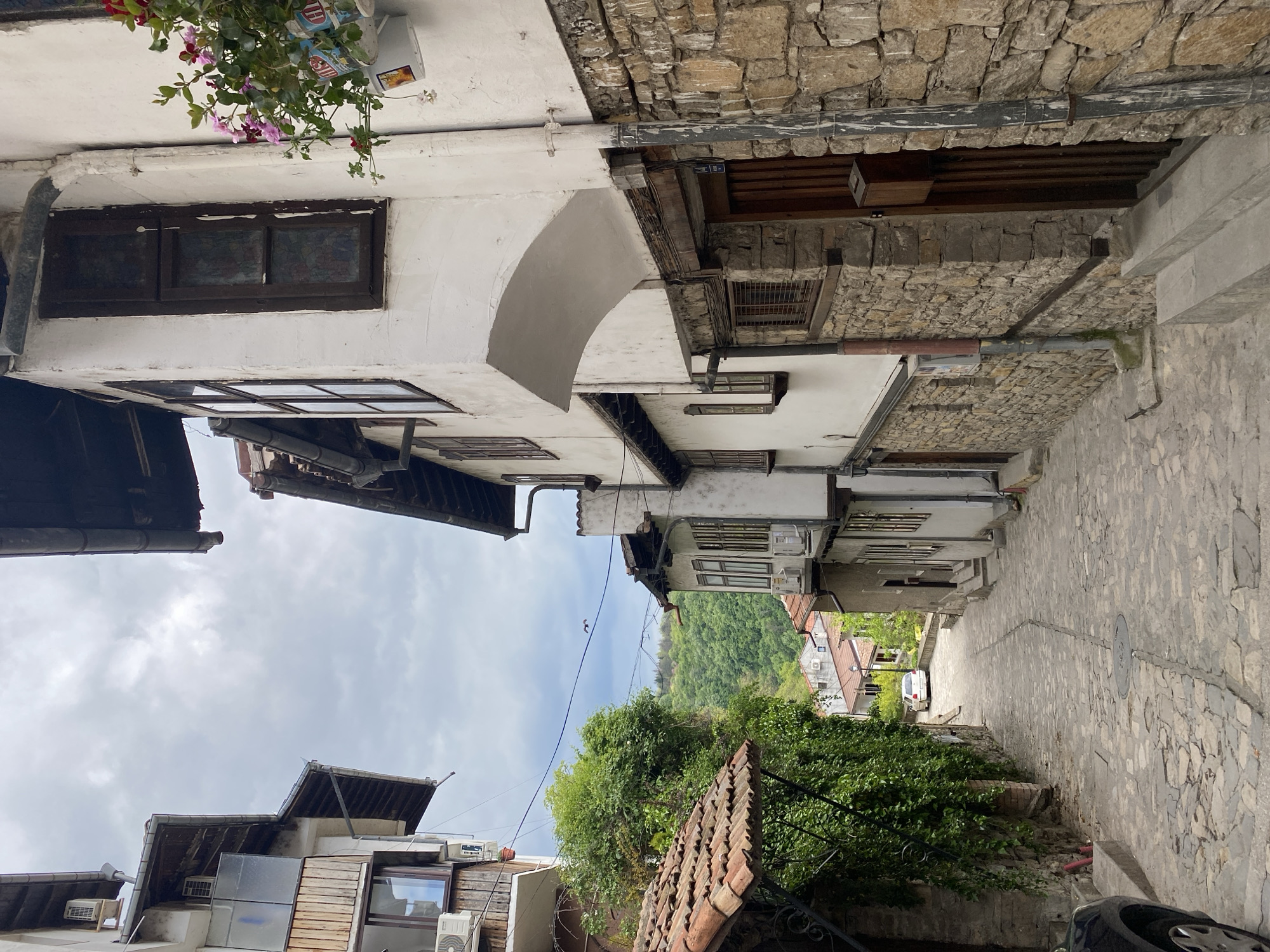 Here is a detailed description of the image:
The image captures a narrow street scene, likely in a historic urban area. The street is paved with stone and appears slightly damp, suggesting recent rain or washing. Buildings line both sides of the street, exhibiting a mix of architectural styles. Some buildings are constructed with stone walls, while others have white-painted facades. Windows of varying sizes and designs punctuate the buildings, some with dark wood frames and others with more modern frames. A few air conditioning units are visible, protruding from the building walls. Above the buildings, a patch of sky is visible, partially obscured by cloud cover. Greenery is also present, with trees and shrubs adding a touch of nature to the urban landscape. A car is partially visible at the end of the street. The overall atmosphere suggests a blend of historical charm and modern functionality.