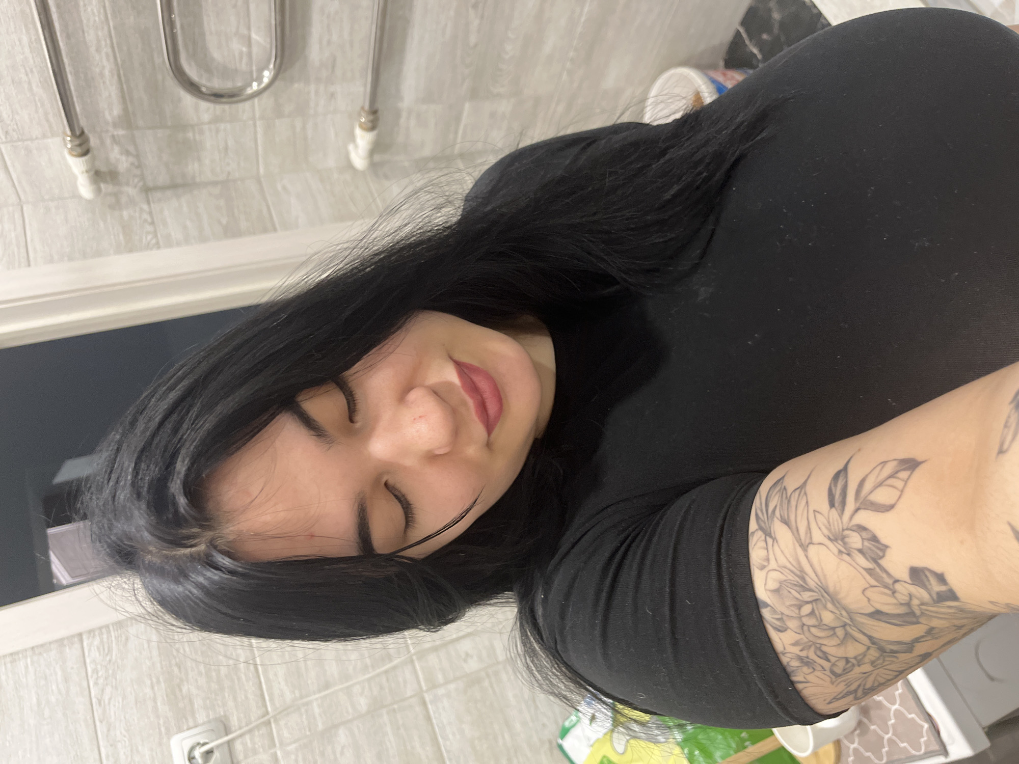 A close-up, self-portrait style photograph depicts a female subject indoors, likely within a bathroom setting. The subject has long, dark hair, with her eyes closed and a slight smile on her lips, which are tinted with pink lipstick. She is wearing a black, short-sleeved top. A large, grayscale floral tattoo is visible on her left upper arm.
The background features light-colored, possibly beige or grey, rectangular wall tiles. To the left, a white electrical outlet with a white cord plugged in is visible on the tiled wall. To the right, a chrome-finished heated towel rail with white components is mounted on the wall. A white door frame or trim is partially visible. In the lower left portion of the image, parts of white fixtures, possibly a sink or toilet, a white ceramic cup, and a green and white item, likely a package, are present. In the lower right, a white plastic bucket with blue and red markings rests on a dark-colored floor. The precise location within the city of Chaikovsky, Russia, cannot be ascertained from the visual evidence.