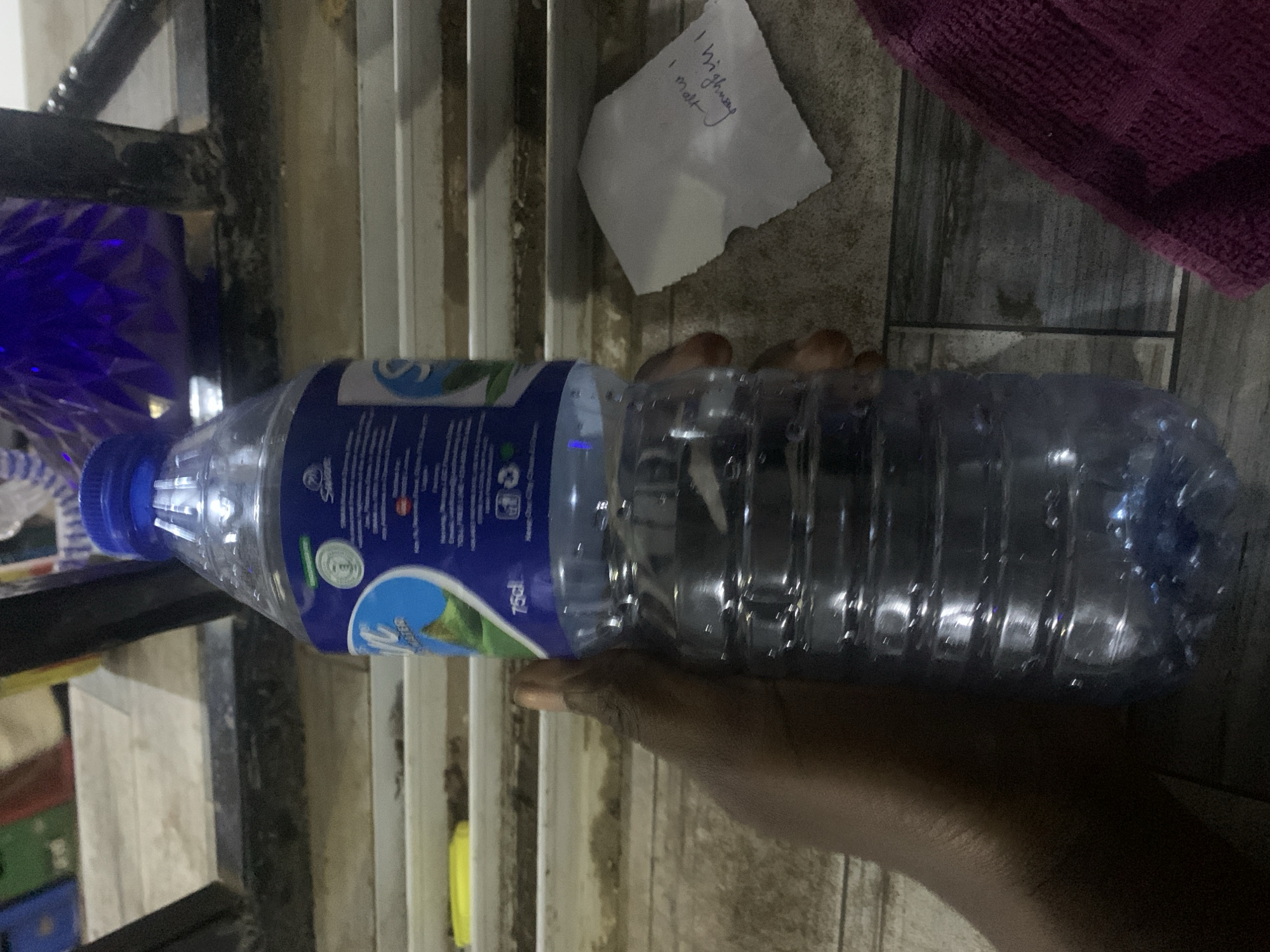 The image presents a dark-skinned hand holding a 75cl plastic bottle of "Sion Water" with a blue cap and label, approximately two-thirds full of clear liquid. The scene is located in New Stadium, Nigeria. To the immediate right of the bottle, on a light-colored surface, rests a small, crumpled piece of white paper with handwritten text, appearing upside down, which includes "I high way I matt." Further to the right and in the foreground, a folded purple textured fabric, consistent with a towel, is partially visible. The background features multiple horizontal light-colored ledges or shelves, with a yellow object positioned on one of the lower ledges to the left. In the upper right background, a prominent blue, faceted object, resembling a decorative container, is partially visible behind a dark, weathered metal structure, possibly a railing or support. The overall environment appears to be an indoor or semi-sheltered area with varied surfaces and objects.