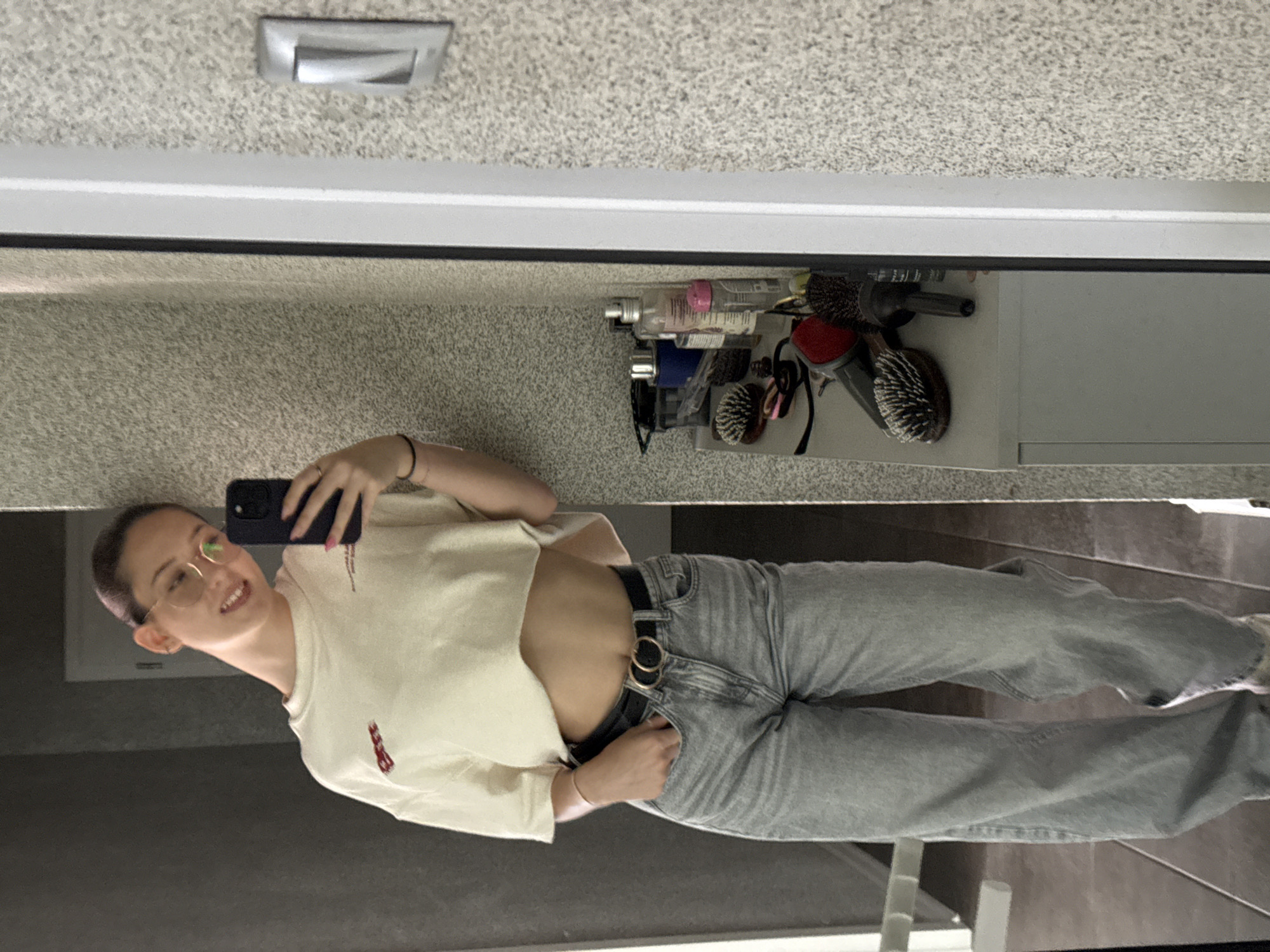 A young woman with glasses and a light-colored crop top is taking a mirror selfie in what appears to be a bathroom in Sofia, Bulgaria. She is wearing light-wash denim jeans and a black belt with a double gold buckle. Her left hand is in her jeans pocket, and she is holding her phone in her right hand to take the picture. Behind her, a bathroom vanity with various toiletries and hairbrushes is visible. The wall has a textured finish.