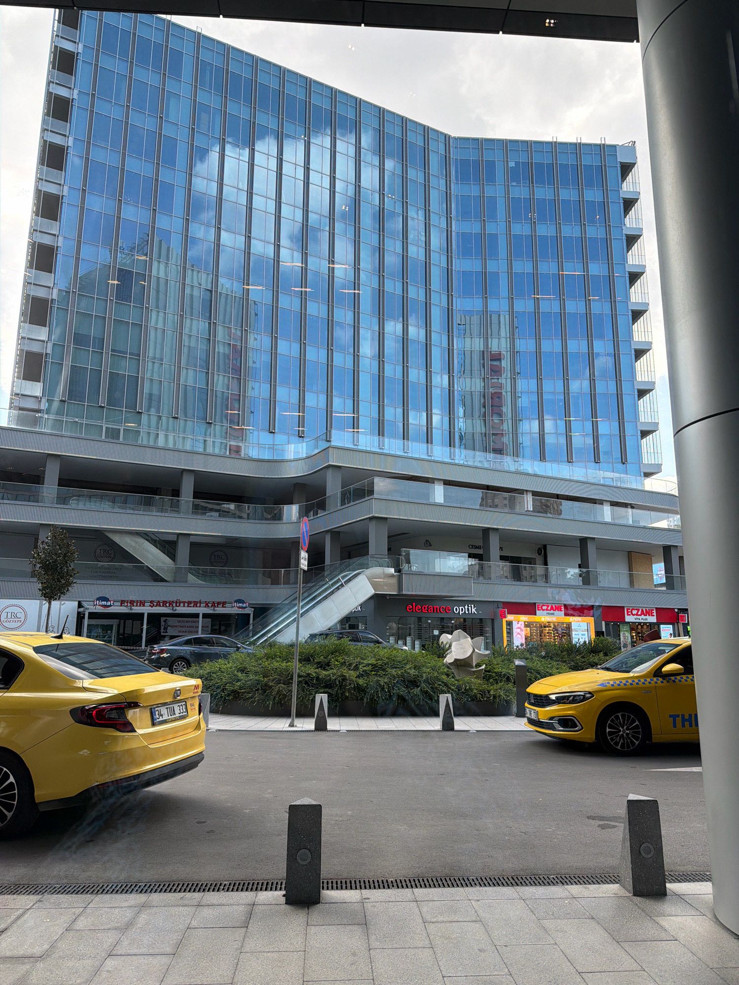 The image captures a street scene in Ataşehir, Turkey, dominated by a modern, multi-story building with a curved facade clad in reflective blue glass. The building's architecture features multiple recessed levels with balconies and walkways. Below the main structure, several retail storefronts are visible, including "elegance optik" and "ECZANE."
In the foreground, two yellow taxis are present. The taxi on the left is a Fiat Tipo with license plate "34 TUA 333" and appears to be emitting exhaust fumes. The taxi on the right is partially visible and also yellow.
The street level is paved with asphalt, and sidewalks are constructed with grey square tiles. Decorative, dark grey bollards are placed along the sidewalk. Green shrubbery and trees are integrated into the urban landscape, softening the concrete and glass environment. The sky is overcast, with visible clouds reflected in the building's glass facade, creating a sense of depth and mirroring the surrounding environment.