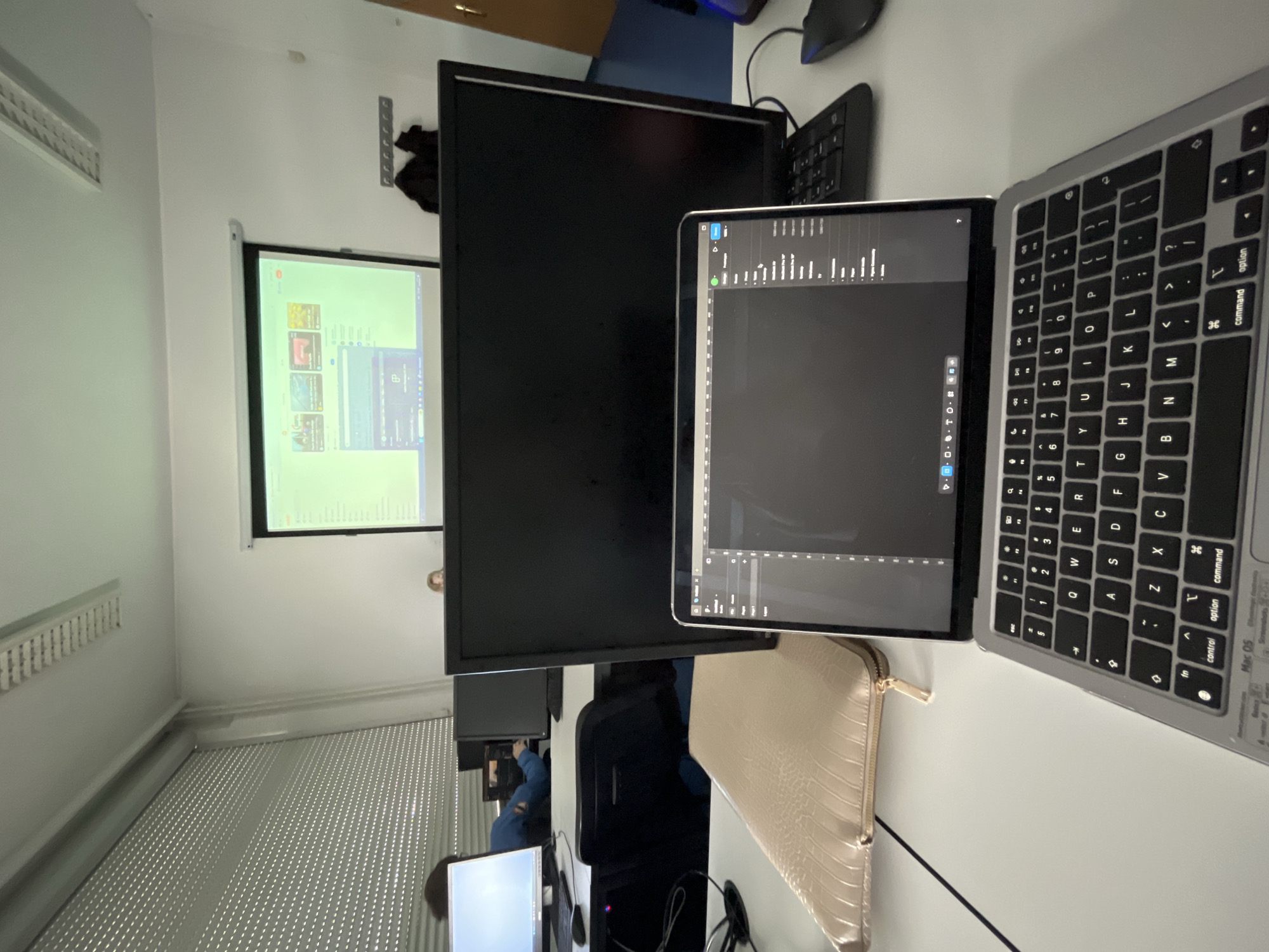 The image is taken indoors, apparently in an office or classroom setting. A laptop, open and displaying some sort of data visualization or programming interface, is prominent in the foreground. To the left of the laptop is a larger monitor, seemingly displaying a blank screen, partially obscuring a projection screen on the wall in the background. The projection screen shows what appears to be a desktop interface, possibly for video conferencing or a shared workspace.
A separate keyboard and mouse are visible, indicating a potential workstation setup. The desk is white. The room has blinds on the window. Other desks and monitors are visible in the background, along with seated individuals suggesting other people are working in the room. The presence of multiple monitors, keyboards, and computer equipment suggests a technology-oriented environment. The location is unknown.