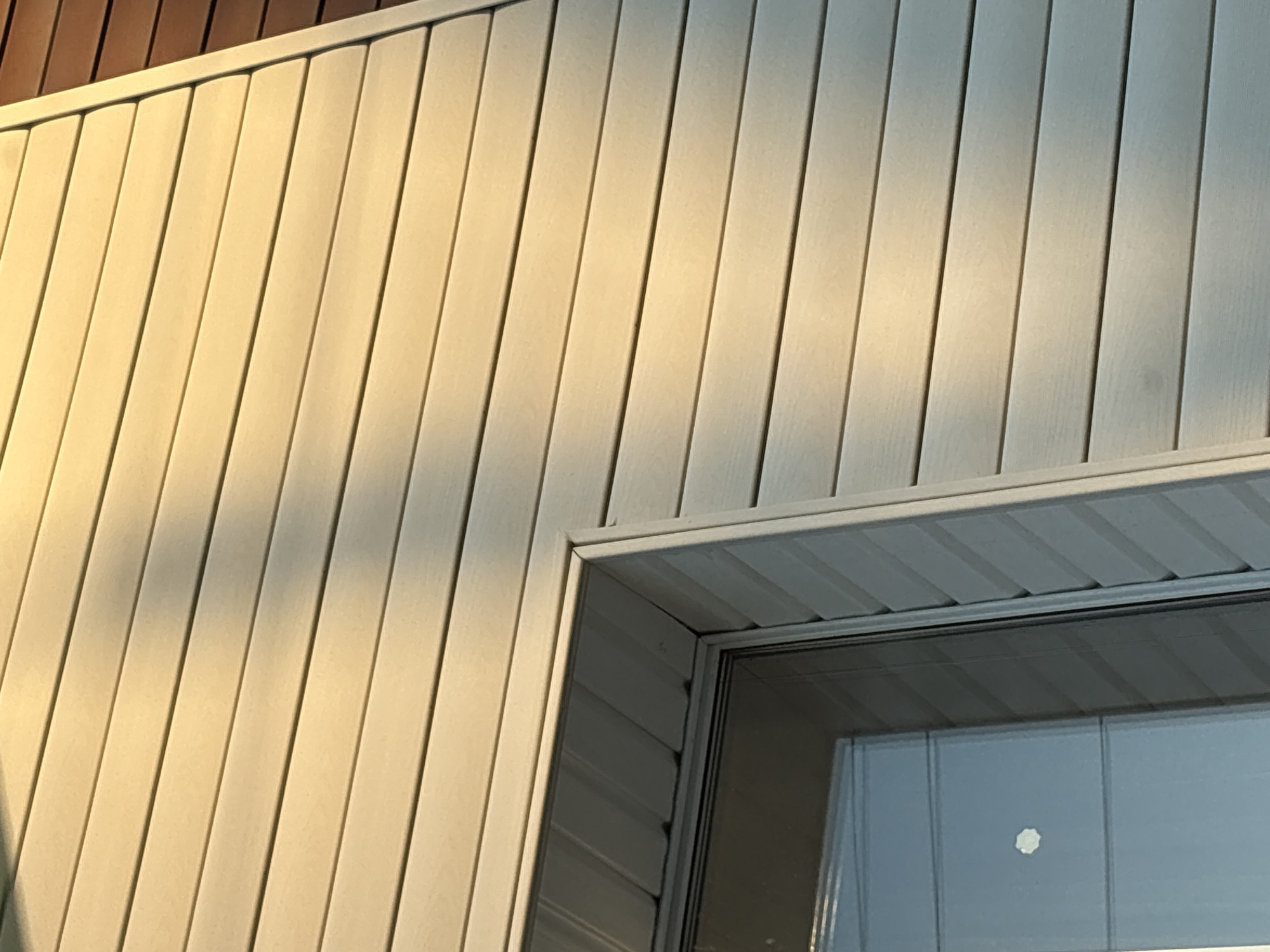 The image shows a close-up of a wall with vertical siding, likely vinyl or similar material, in a light beige or off-white color. The siding is oriented vertically, and there are shadows and highlights across the surface, indicating a light source is present. The siding appears to be part of a building exterior. In the lower right corner of the image is a window with a white frame. The window appears to be closed and reflects light. Part of the structure also looks like brown wood. Based on the information available, the location might be in Прикамский, Russia.