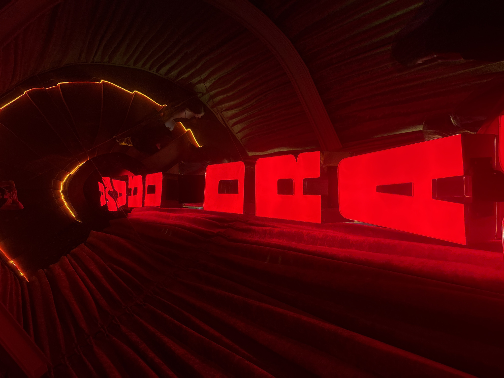 The image depicts a red-lit, tunnel-like installation with large, red letters spelling out a word. The tunnel is lined with textured material, possibly fabric or corrugated metal, with yellow neon lights accentuating the curved edges of the tunnel opening. The letters appear to be illuminated from within, casting a bright red glow. At the far end of the tunnel, the letters diminish in size due to perspective, creating a sense of depth. The overall environment is dark, with the red lights dominating the scene. While specific details are limited, the scene suggests an artistic or theatrical installation, potentially located indoors. Without further information, the exact location within Mexico City cannot be determined.