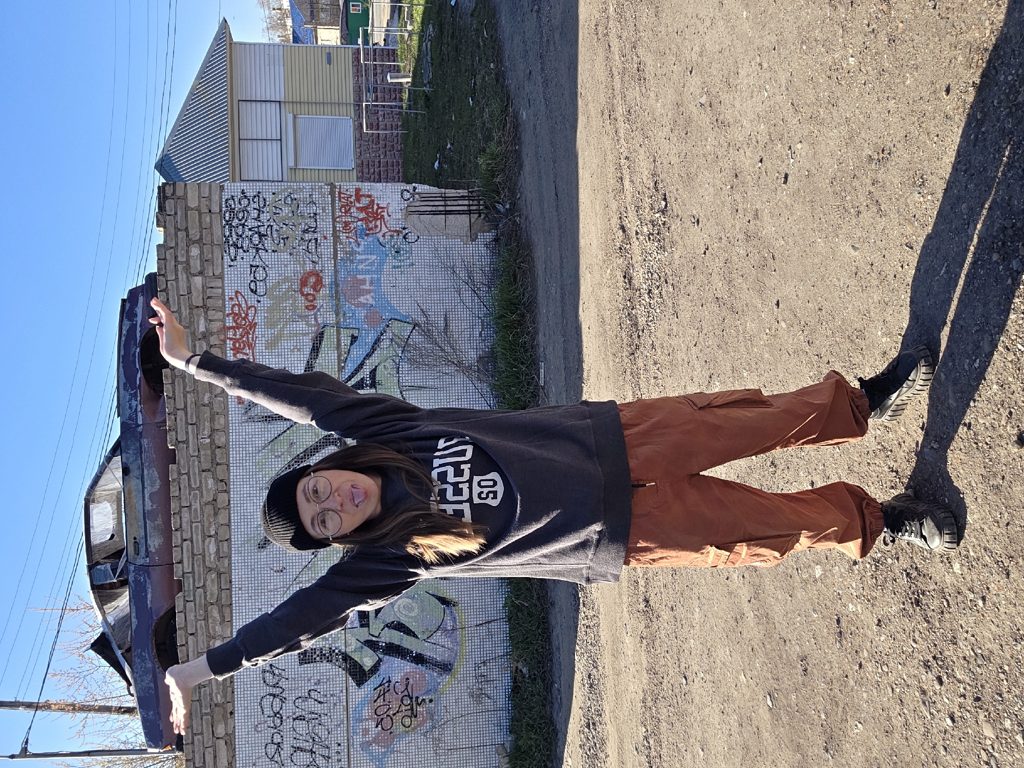 A woman stands with arms outstretched beside a graffitied wall and a burned-out car in Chaikovsky, Russia. She wears a black beanie, round glasses, a dark sweatshirt with white lettering, and orange cargo pants with black shoes. Her tongue is sticking out. The wall is decorated with graffiti and mosaic tiles. A sandy or gravelly ground stretches to the right. Beyond the wall, a two-story building with a metal roof and light-colored siding is visible, along with some foliage. The sky is blue. The scene appears to be outdoors, possibly in a neglected or industrial area of Chaikovsky, Russia.