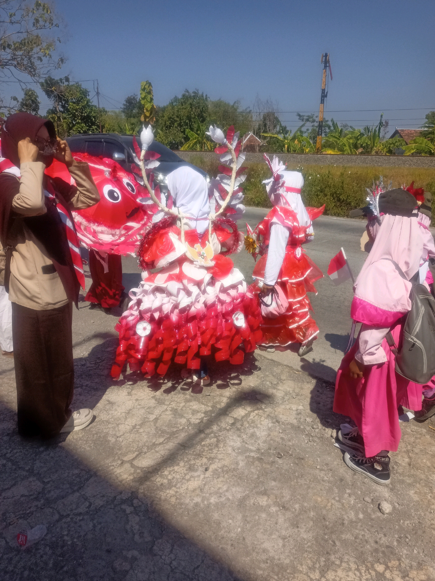 On a sunny day in Sumberlawang, Indonesia, a group of people in festive red and white costumes are gathered outdoors. The individuals appear to be participating in a parade or event, with some wearing elaborate costumes that resemble red and white flowers or abstract art. One costume in the foreground is particularly striking, made of layered red and white materials that hang down like petals. Another person on the left is wearing a brown uniform and a hijab, adjusting their glasses. In the background, a car is decorated with a large red, cartoonish face with white eyes. Other individuals in the image are also wearing similar red and white attire, some with head coverings and some with decorative headpieces. A small Indonesian flag on a stick is visible, held by one of the participants. The ground is paved with a rough, gray surface, and shadows are cast by the sun. Beyond the immediate gathering, trees and vegetation line the background, with a railway track and a utility pole also visible, suggesting the location is near a rural or semi-rural area.