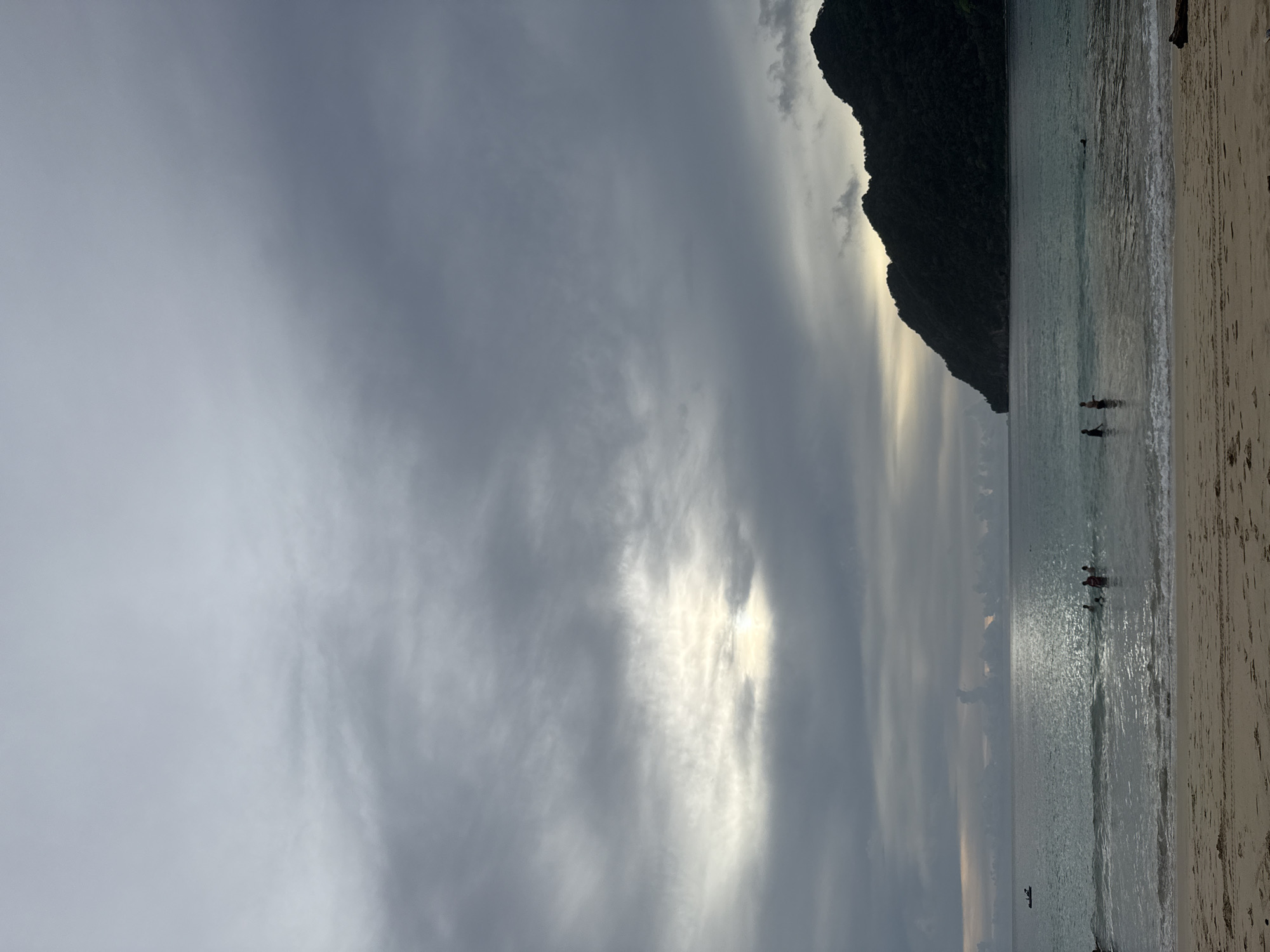 The image depicts a serene beach scene in Meunasahbale, Indonesia, likely during the late afternoon or early evening. The sky is filled with dramatic, swirling grey clouds, with patches of sunlight breaking through, suggesting the possibility of an impending sunset or a break in the weather. A dark, silhouetted mountain juts out from the right side of the frame, its peak covered in vegetation and its base meeting the water.
The calm, shallow ocean reflects the muted light of the sky. Several people are visible in the water, wading or swimming near the shore, their figures small against the vastness of the sea and sky. A small, dark boat is also present on the water further out. The sandy beach occupies the rightmost portion of the image, appearing relatively empty except for some subtle footprints and a piece of driftwood. The overall mood is one of tranquility and natural beauty, with the dramatic sky adding a touch of atmospheric intensity to the peaceful coastal setting. No text is visible in the image.