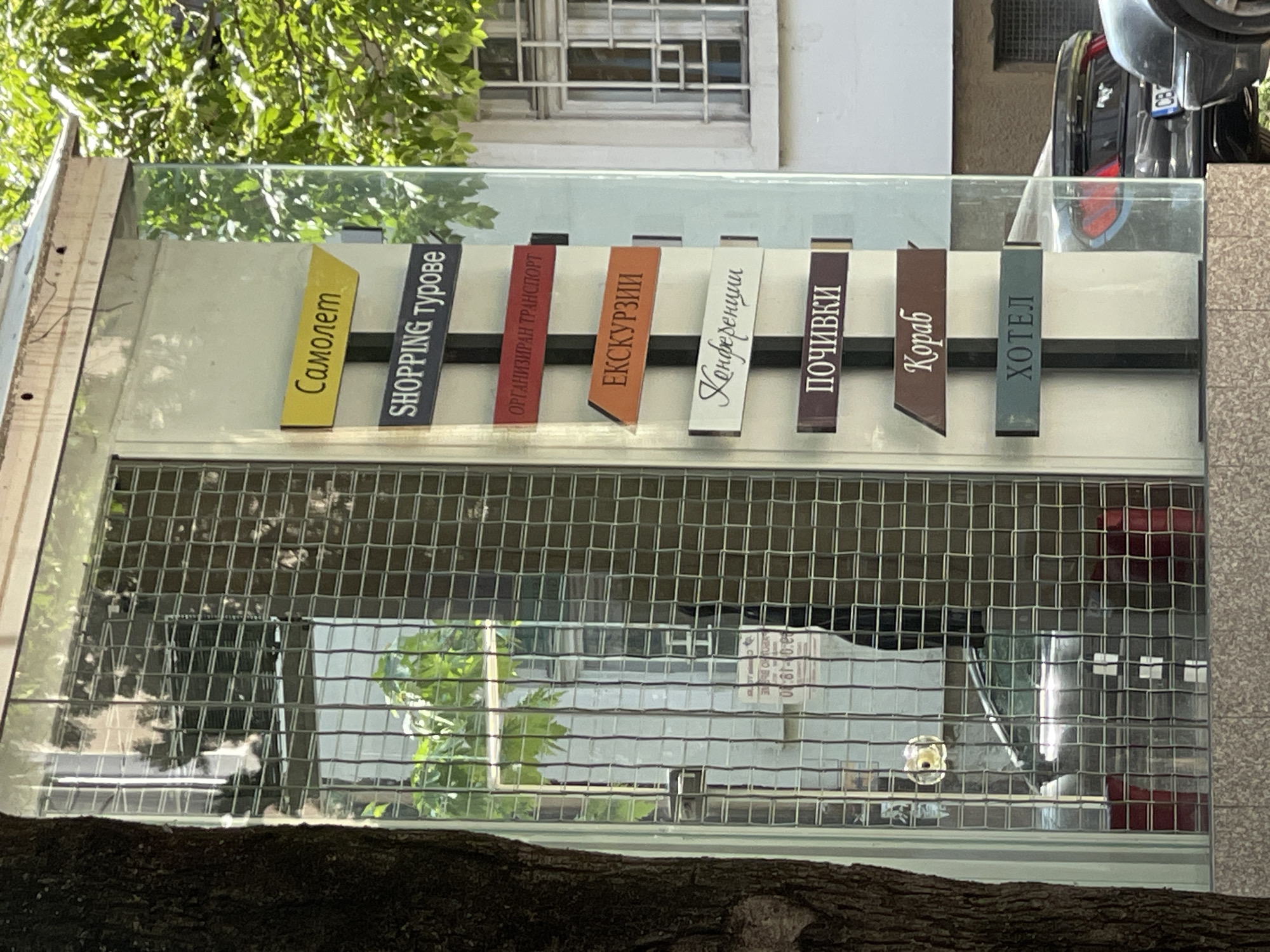 The image shows a storefront in Sofia, Bulgaria, with a sign listing various services in Cyrillic: "самолет" (airplane), "shopping турове" (shopping tours), "организиран транспорт" (organized transport), "екскурзии" (excursions), "конференции" (conferences), "почивки" (vacations), "кораб" (ship), "хотел" (hotel). The sign is made of vertically-oriented color-coded panels. The store window has a wire mesh covering. A tree branch partially obscures the lower portion of the view. The upper portion of the image includes part of a building exterior. A black vehicle is partially visible in the upper right corner.