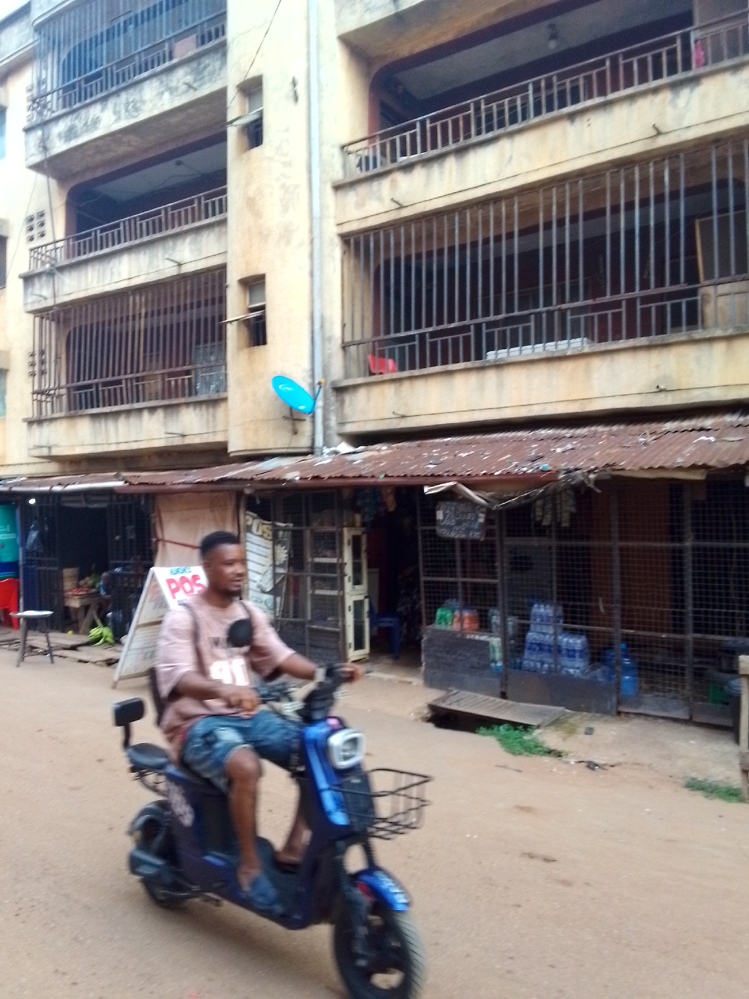 A man rides a blue scooter on a dirt road in Onitsha, Nigeria. The scooter has a basket on the front and a seat back. The man wears a pink t-shirt, shorts, and sandals. Behind him are shops with metal security bars. Some of the shops have rusty corrugated metal roofs. Bottles of water are displayed in one shop. Behind the shops is a multi-story concrete building with barred windows and balconies. A blue satellite dish is attached to the building. A POS sign is next to one of the shops.