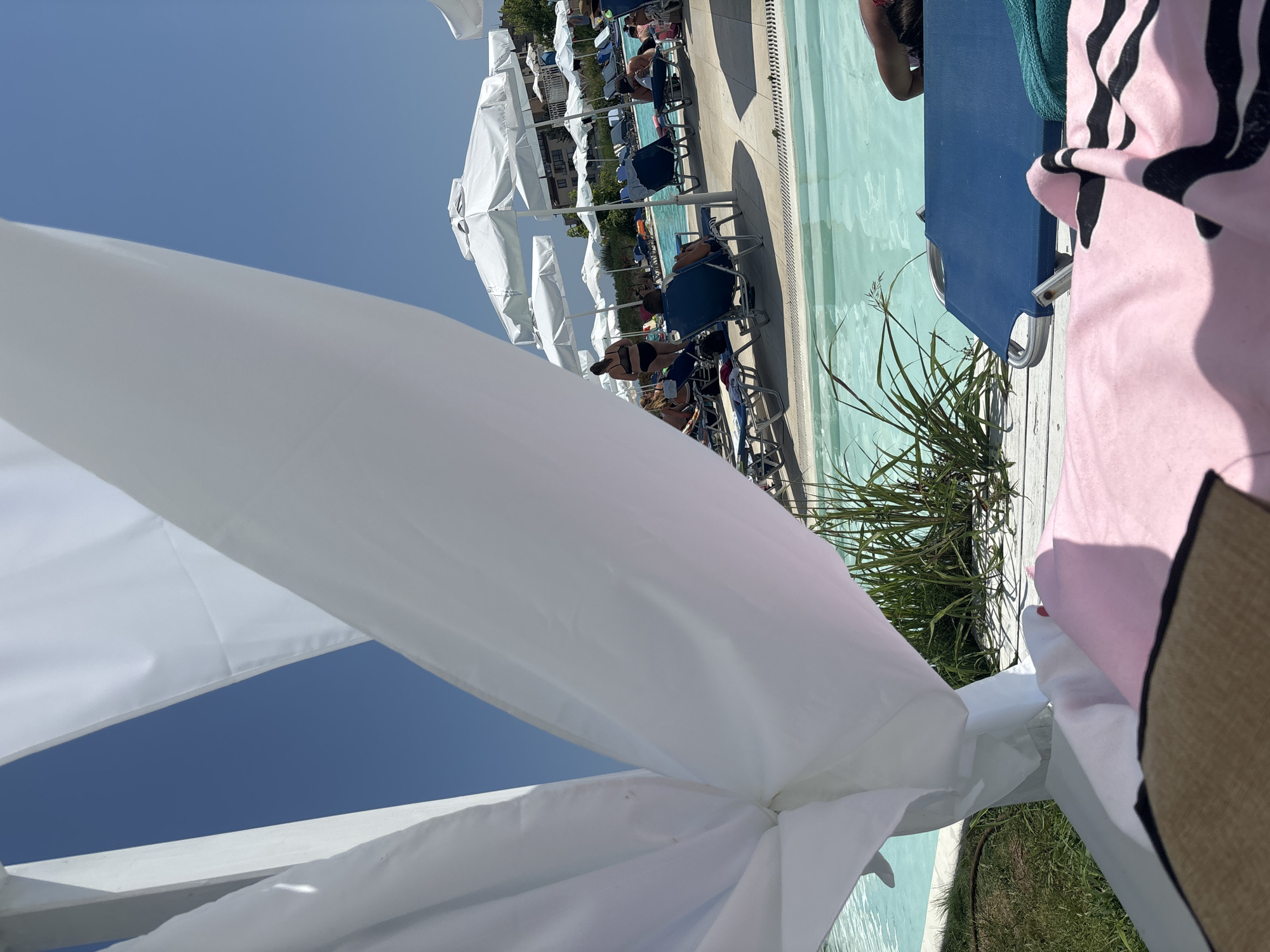 In Byala, Bulgaria, a sunny day unfolds at a resort, framed by flowing white fabric. In the foreground, a pink towel with black stripes and a woven hat lie on a wooden deck. Beyond, lounge chairs and umbrellas line a sparkling turquoise pool. People relax on chairs, some reading, others chatting, while a woman in a black bikini stands near the water's edge. Lush green grass and tall reeds add to the vibrant scene, with apartment buildings visible in the distance. The clear blue sky overhead completes this idyllic summer setting.