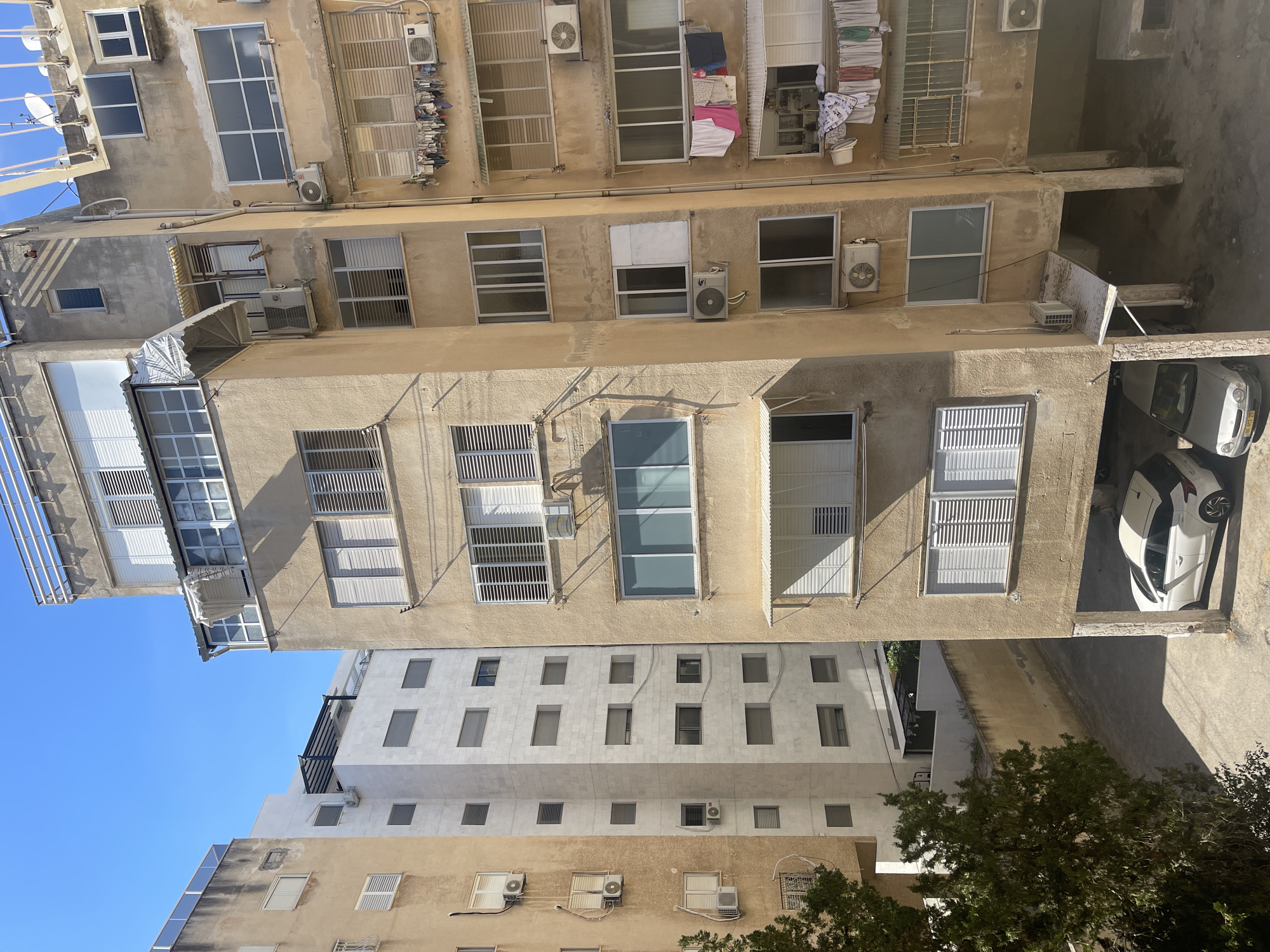 This media file captures an outdoor scene of apartment buildings in Netanya, Israel, likely on a clear, sunny day. The perspective is from a higher vantage point, looking down and across the buildings. Multiple building facades are visible, characterized by light-colored, weathered plaster walls and numerous windows, many with horizontal slatted blinds.
Several objects are noticeable, including air conditioning units mounted on the exterior walls of various apartments. Laundry is also visible, hanging on clotheslines outside some windows, suggesting a domestic setting. In the lower right of the frame, a white car is parked in an open-air parking area, partially visible.
The architecture suggests an urban or suburban residential environment. The clear blue sky in the upper left portion of the image indicates fair weather and likely daytime, though the specific time is not determinable. There are no people clearly visible in the image, and the overall impression is one of a quiet, everyday scene. No text is discernable in the image.