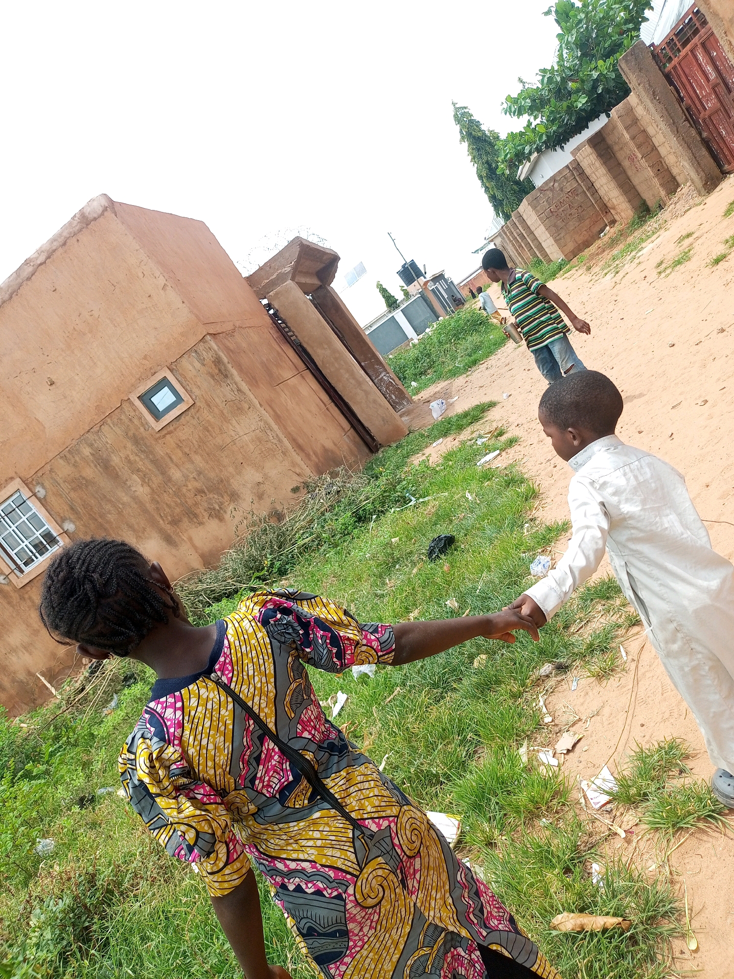The image depicts a sandy, unpaved street in Tempure, Nigeria. In the foreground, a young person with braided hair, wearing a brightly colored yellow, pink, and gray patterned dress, is holding hands with a younger child dressed in a white robe. The child in the white robe is extending their right hand towards the person in the patterned dress. Behind them, a slightly older child in a striped t-shirt and jeans walks away from the camera, holding a metallic cup. The scene is bordered by overgrown green grass and a low-rise, tan-colored building with a square window. Further in the background, a fence made of brick and metal is visible, with trees and more buildings beyond. The sky is overcast and white. The ground is littered with small pieces of white paper and some darker debris.