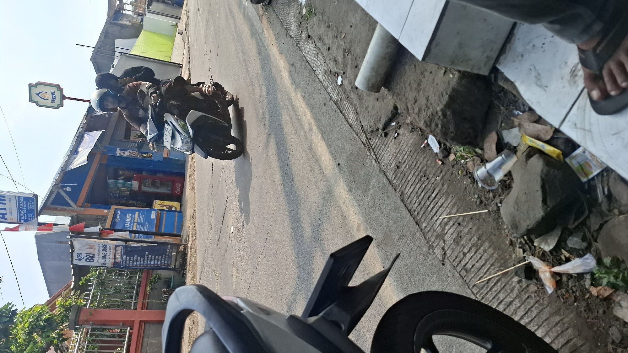 The image captures a street scene in Cilaku, Indonesia. In the foreground, a motorcycle is parked, its rear wheel and fender visible. The motorcycle is a modern, light-colored scooter. The road surface is asphalt, with cracks and a textured edge along the sidewalk. A sidewalk, constructed with concrete slabs, is littered with debris, including plastic bags, wrappers, a discarded plastic cup, and small wooden sticks. Several rocks are also scattered on the ground.
In the midground, a man wearing a helmet is riding a scooter, carrying a passenger also wearing a helmet. They are on the road, heading away from the viewer. The scooter appears to be a similar model to the one parked in the foreground.
In the background, there is a small building with a blue and orange facade, likely a shop. Signs are visible on the building and a nearby pole. One sign, vertically oriented and red and white, appears to be a flag. Another sign, blue and white, displays text related to "BRI" and "UJANG JUBAEDI". A prominent "ATM" sign is also visible at the top of a pole. Further in the background, there is a green wall and some trees. The sky is clear and bright, indicating daylight. The overall impression is a typical street scene in a developed area of Indonesia, with elements of commerce and transportation.