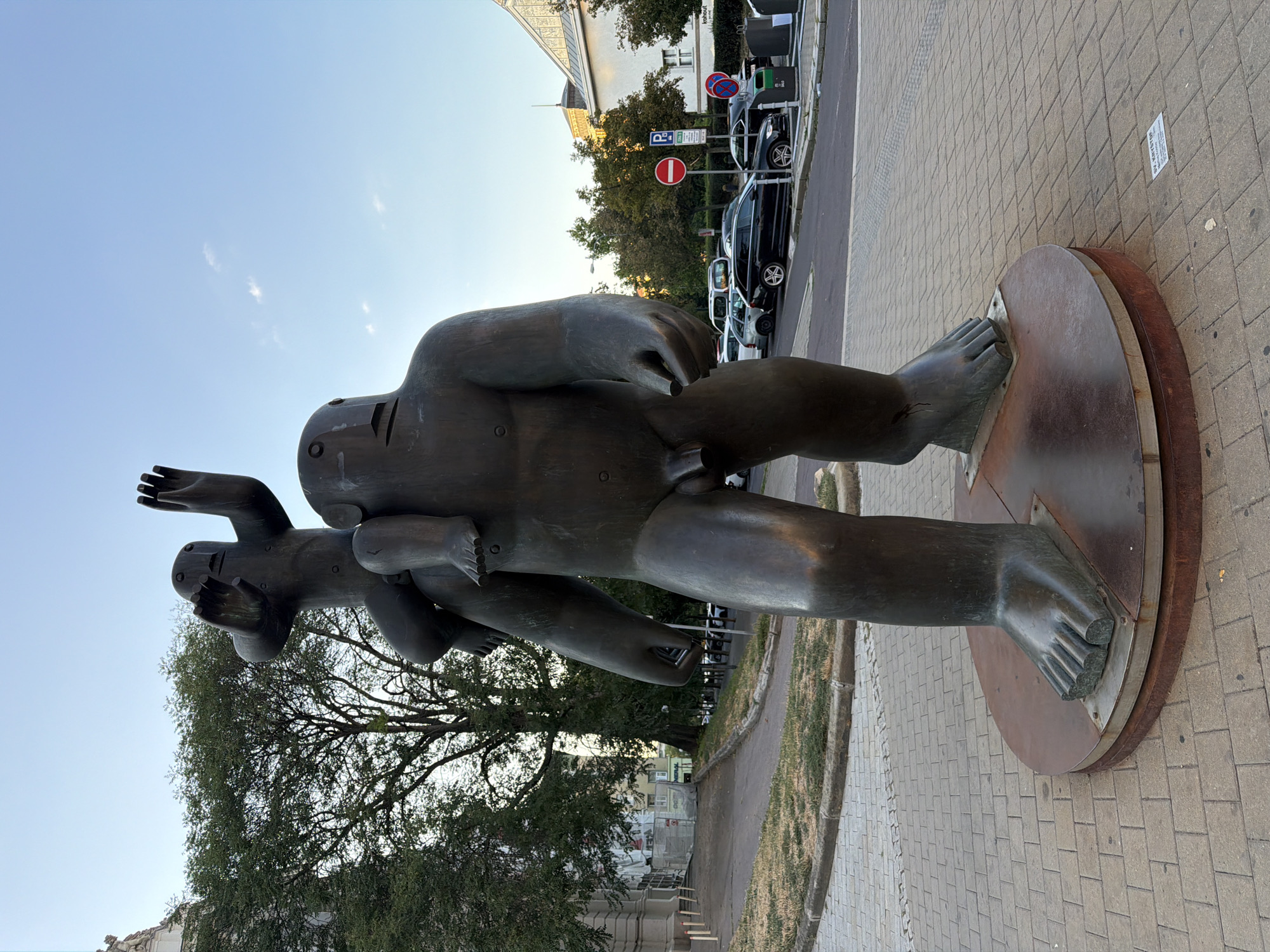 A large, dark bronze statue of two figures stands on a circular metal base, which is set on a patterned brick sidewalk. The lower, larger figure is a male with a large torso and legs, standing upright. He has a simple, rectangular face with circular eyes and a horizontal slit for a mouth. His arms are down at his sides. Perched on his shoulders is a smaller, childlike figure, also facing forward. This figure has its arms raised outwards and slightly upwards, with its hands open and palms facing forward. It has a similar stylized face to the larger figure. The larger figure's left hand is partially visible, holding something small and rectangular. The statue is located in front of a building with a light-colored facade and large windows. Behind the statue, a street is visible with parked cars and traffic signs. Trees with green foliage line the sidewalk and street. The sky above is clear and blue with a few wispy clouds. The image is taken in daylight, likely late afternoon or early evening due to the soft lighting. The statue is located in Brno, Czechia, specifically at Moravské náměstí (Moravian Square).