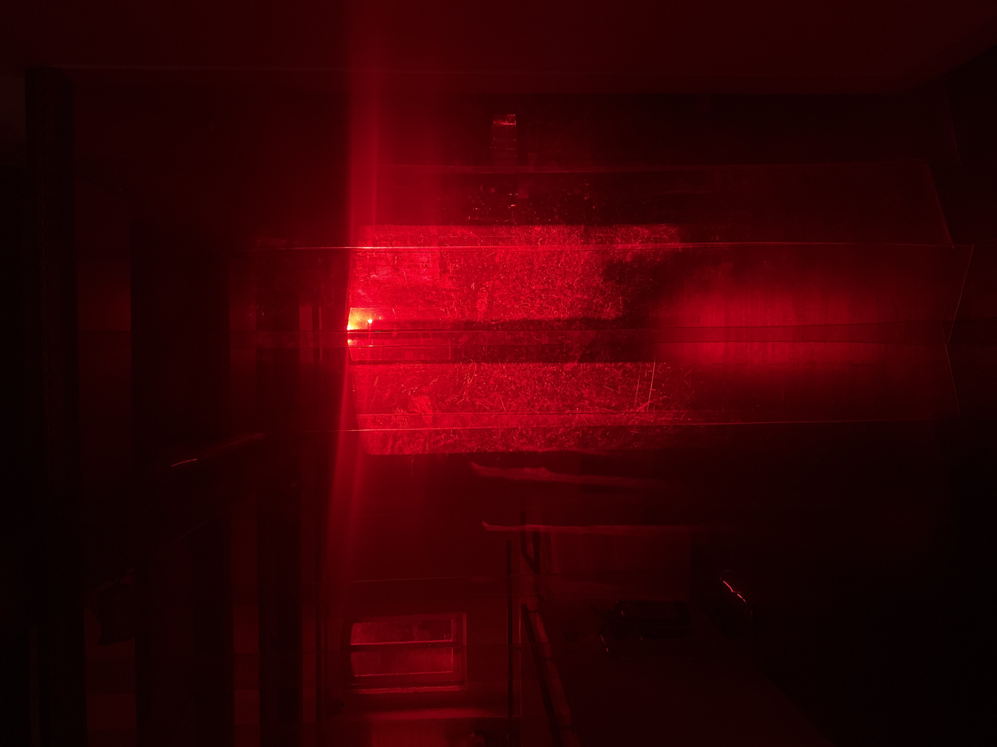 The image presents a dark, indoor scene heavily dominated by an intense red light, creating a mysterious and somewhat industrial atmosphere. No people are visible. The central focus is a series of horizontal, layered structures or platforms, possibly made of translucent plastic or glass, which are brightly illuminated by a strong red light source seemingly positioned to the left.
These illuminated surfaces display a textured appearance, suggesting dust, scratches, or condensation, which diffuses the red light and creates a mottled, almost web-like pattern across them. The light casts deep shadows, obscuring details in the surrounding areas and enhancing the sense of depth. In the lower-left quadrant, a smaller, dark structure features a rectangular opening with horizontal bars, resembling a window or vent, which reflects the red light. Faint outlines of what might be railings or structural elements are discernible in the darker regions.
The pervasive red lighting suggests an artificially lit environment, likely at night or within a specialized space. There is no clear activity taking place. While the image itself lacks specific locational cues, the context provided indicates the scene is located in London, United Kingdom. The overall impression is one of an abstract visual display, possibly an art installation, a darkroom, or a uniquely lit interior space. No text is visible within the image.