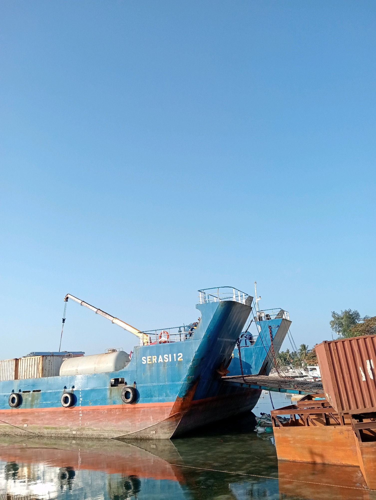In the city of Pagerungan Kecil, Indonesia, two large blue cargo ships are docked in calm water under a clear blue sky. The ship in the foreground, named "SERASI 12," displays its name in gold lettering. It is laden with containers and has a grey cylindrical tank on its deck. Several black tires are mounted along its side. A crane extends from the ship towards the left side of the frame. A ramp connects the "SERASI 12" to another docked ship behind it, which also appears to be blue and loaded with containers. To the right, a rust-colored shipping container is partially visible. The water reflects the ships and the sky. The scene is static, suggesting the ships are either docked or waiting. No people are clearly visible in the frame. The environment appears to be a port or harbor.
