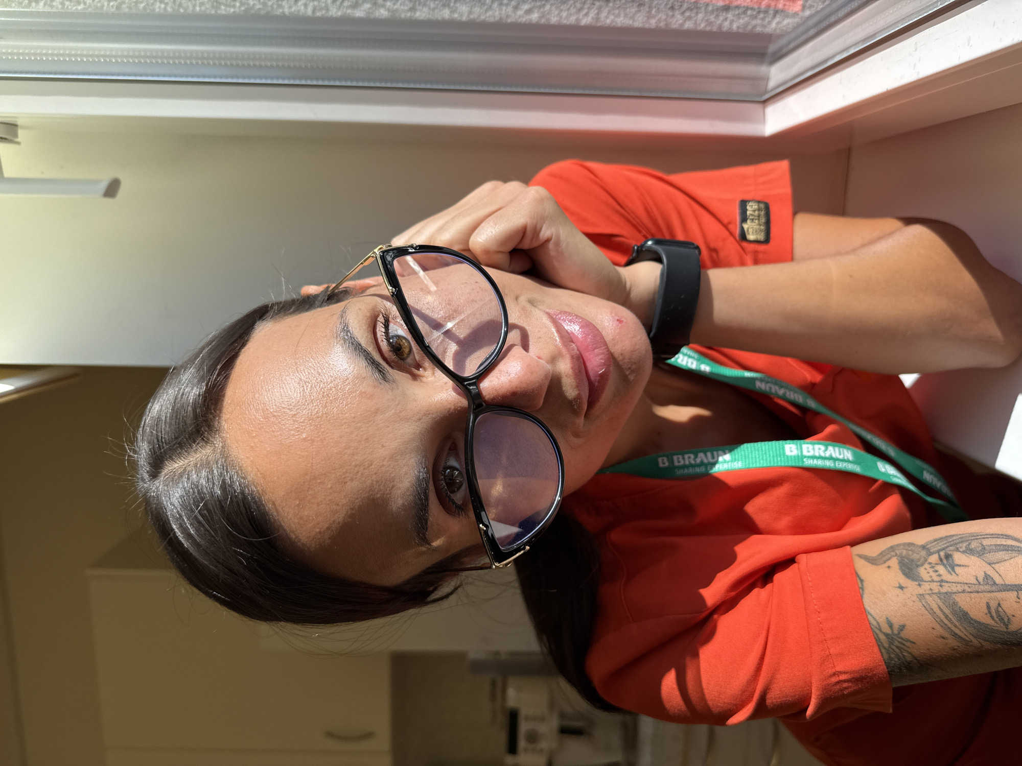 The image shows a medium shot of a woman with dark hair and brown eyes wearing black-rimmed glasses. She is wearing an orange t-shirt with a green lanyard that says "B Braun Sharing Expertise." She has a tattoo on her left arm. The woman is leaning on her hand, with her face turned towards the camera. Sunlight is casting shadows on her face and the background. The background appears to be an indoor setting, possibly a room with cabinets and a window. There is no specific indication of the city of Pleven, Bulgaria in the image itself.
