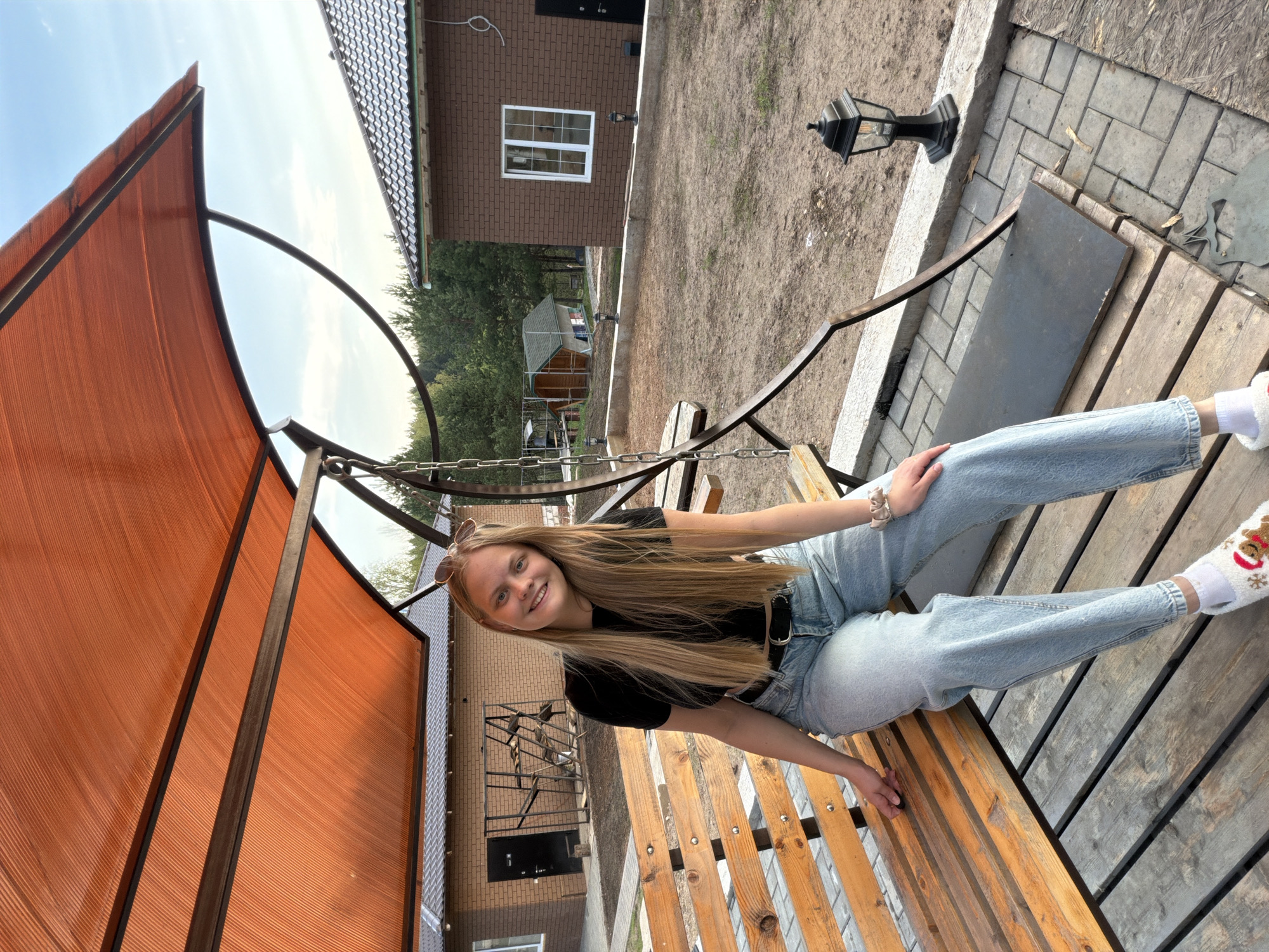 A young woman is sitting on a wooden swing in what appears to be a yard in Kamennoye, Russia. She has long blonde hair, wears sunglasses perched on her head, a black t-shirt, jeans, and white socks with a reindeer design. The swing is under a reddish-orange structure that provides shade. Behind her are buildings, including a brick house with white-framed windows, and a glimpse of a smaller wooden structure. A black lamppost stands on a paved area near the swing. The ground is a mix of dirt and paved surfaces.
