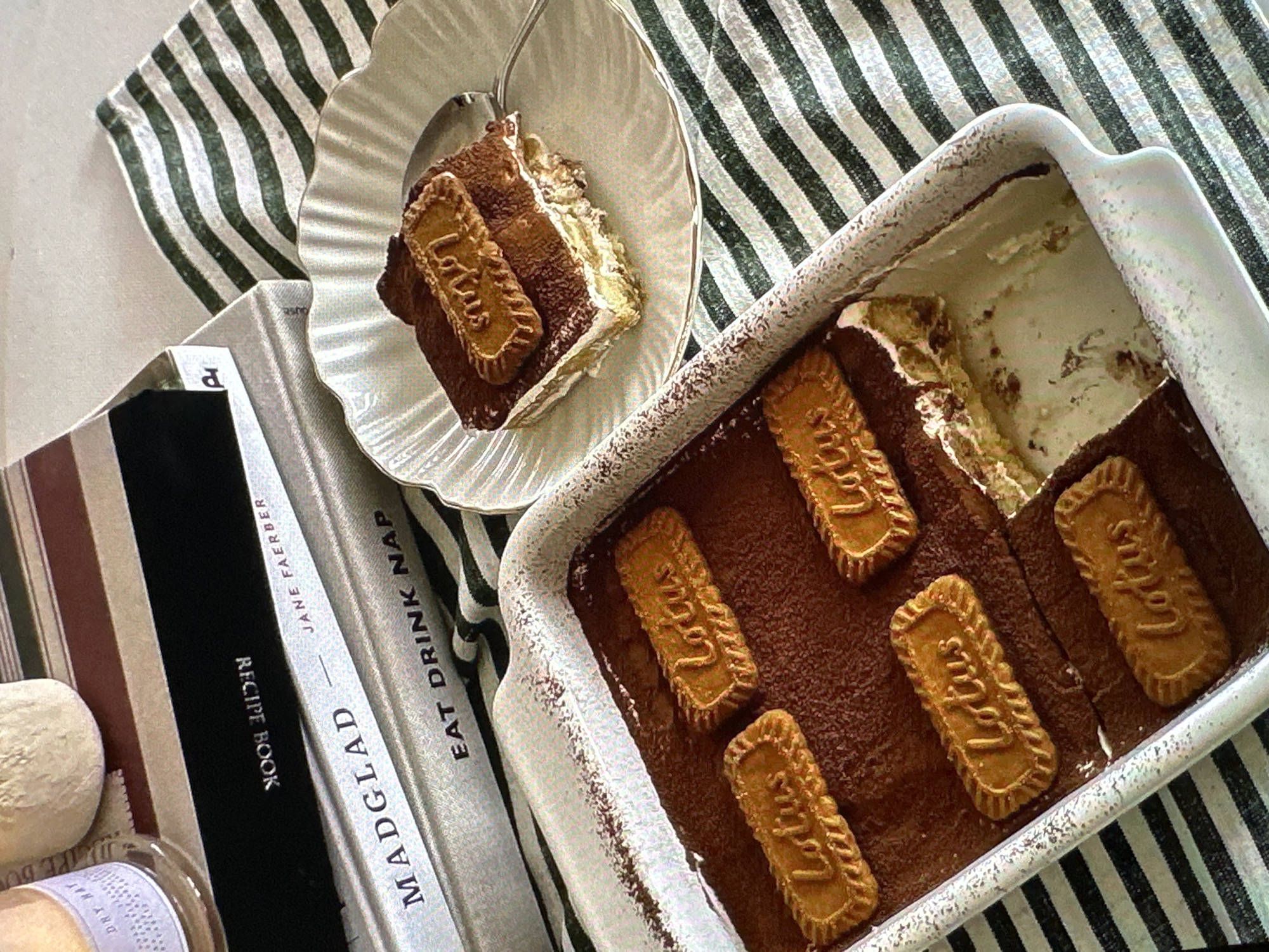 This image shows a portion of Lotus Biscoff tiramisu, on a striped tablecloth, alongside cookbooks and other objects. A piece of the tiramisu is on a decorative plate to the left of the baking dish that holds the rest of the dessert. The top of the tiramisu is dusted with cocoa powder and decorated with Lotus Biscoff cookies. The cookbooks include titles like "MADGLAD," "JANE FAERBER RECIPE BOOK," and "EAT DRINK NAP." A ball of dough or a similar object sits to the left of the cookbooks, and a glass jar is partially visible at the bottom left corner. The location is not specified, so this cannot be determined.
