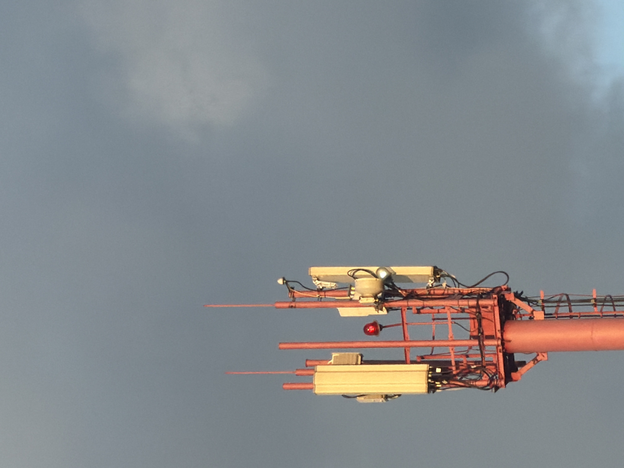 A tall, red cell tower stands against a muted, cloudy sky. The tower has a cylindrical base and a lattice-like structure at its top, adorned with several antennas. One antenna, a prominent rectangular unit, is a pale yellow and has a textured surface. Another, a smaller white unit, is mounted higher up. A bright red, round light is visible on the tower's structure, and a white fixture with a lens is also present. The sky is a uniform gray, with a hint of lighter cloud wisps on the right side. The overall scene is static and industrial.