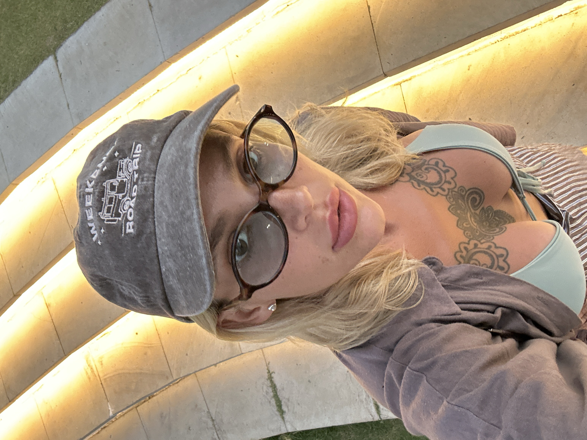 A close-up selfie of a blonde woman wearing glasses and a grey baseball cap is taken outdoors on a set of illuminated concrete stairs. The woman has a tattoo on her chest and is wearing a light blue bikini top. The lighting suggests it is dusk or evening. The location is Kuta Selatan, Indonesia.