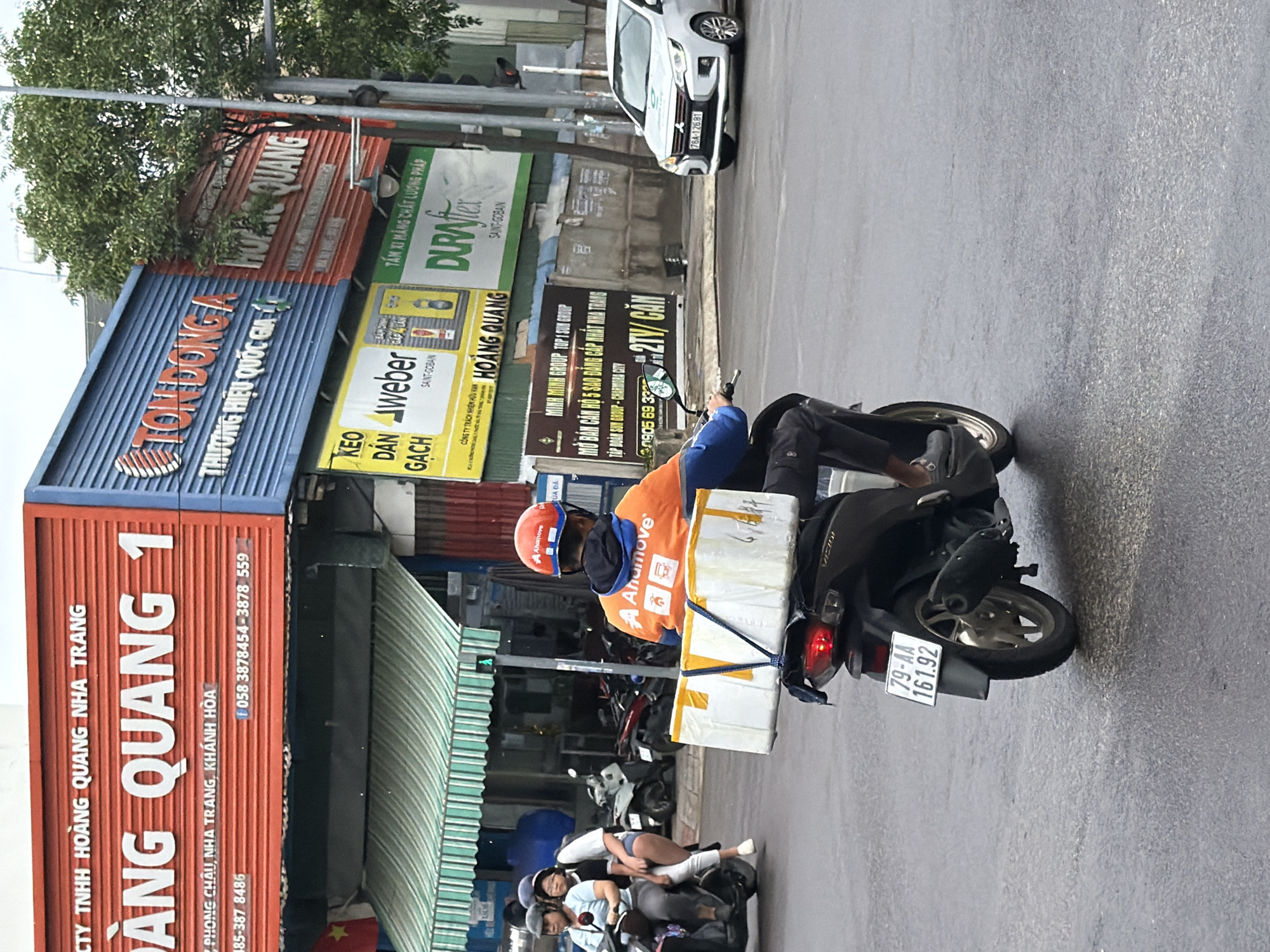 In Nha Trang, Vietnam, a delivery driver on a black motorcycle, identified by the license plate "79-AA 161.92", transports a large white box secured to the rear of the vehicle. The driver wears an orange and blue jacket and a blue helmet with "Anamove" branding. In the background, a white car with license plate "76A-126.81" drives on the wet street. Above the street, a multi-story building displays various advertisements. One prominent sign, in red and blue, reads "HOÀNG QUANG" and "TON DONG A". Another sign is for "DURAFlex". A green and white striped awning extends from the building, and a traffic light is visible. To the left, two women on a scooter are also visible. The overall scene depicts a typical street view in an urban environment with commercial activity and ongoing traffic.