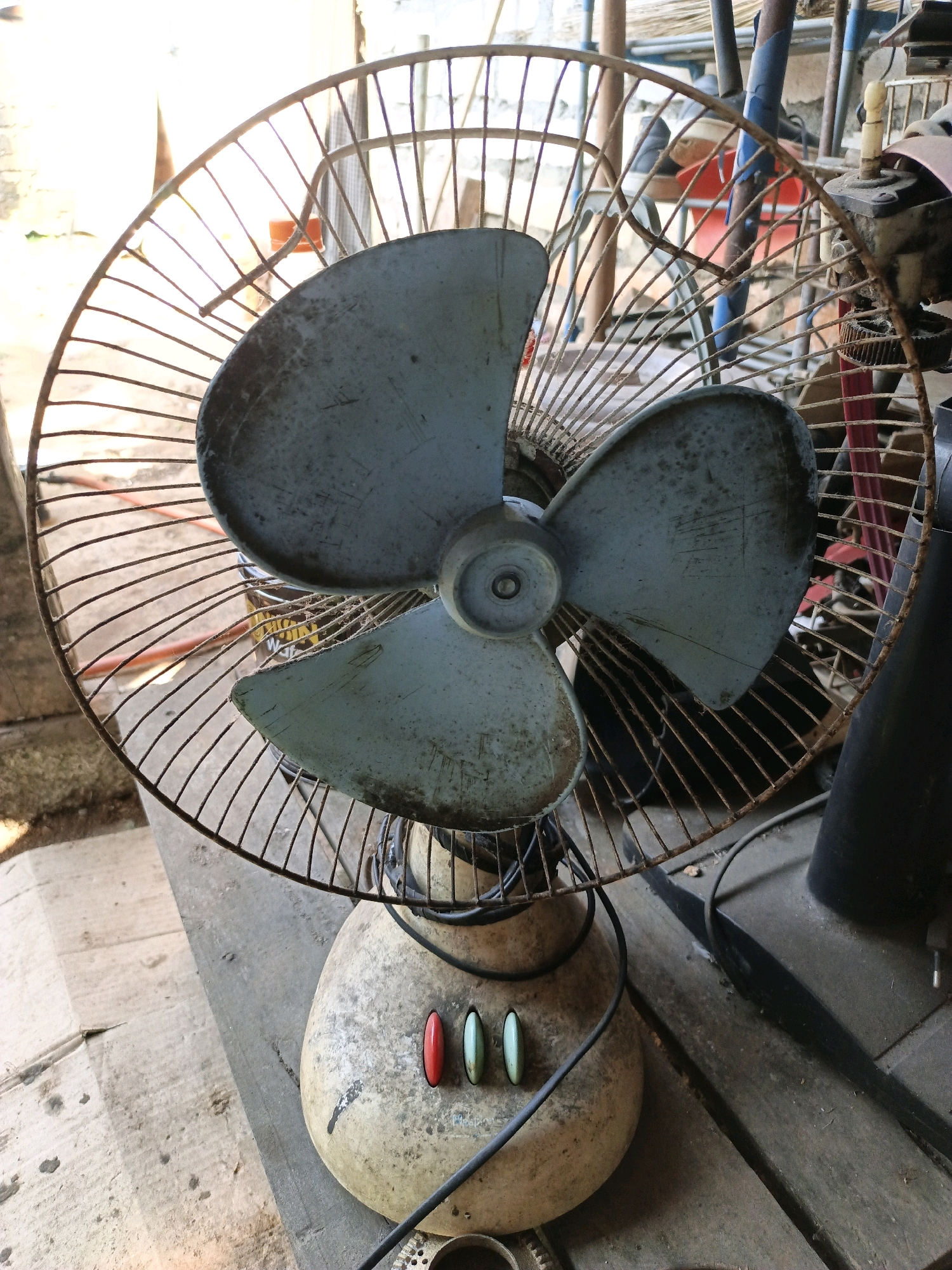 The image depicts a close-up, eye-level view of an old, dusty electric fan. The fan is made of metal and plastic and appears to be in disrepair. Its blades are a faded blue, and the protective grill is rusty. The fan rests on a wooden surface, possibly a table or a floor, with other objects visible in the background that suggest an outdoor or storage setting. Three colored buttons – red, green, and blue – are visible on the fan's base, but their function is unclear. The lighting suggests it might be daytime, with some sunlight filtering in. The location is identified as Keleyan, Indonesia.