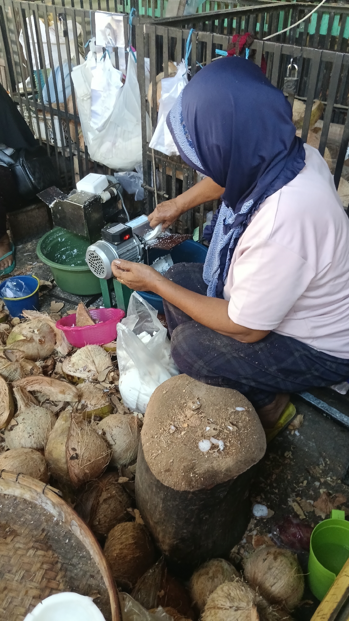 An outdoor scene depicts a woman grating coconuts in a semi-urban environment in Purwokerto, Indonesia. The woman, wearing a blue headscarf and a light pink shirt, is seated on the ground, actively using an electric coconut grater. Piles of coconuts, both whole and halved, surround her, along with various containers, including a green basin filled with shredded coconut, a pink bowl, and a blue bucket. A large wooden chopping block is positioned in front of her, also covered in coconut debris. In the background, a metal fence with hanging white plastic bags creates a visual barrier. The ground appears to be dirt and debris, indicative of an outdoor marketplace or processing area.