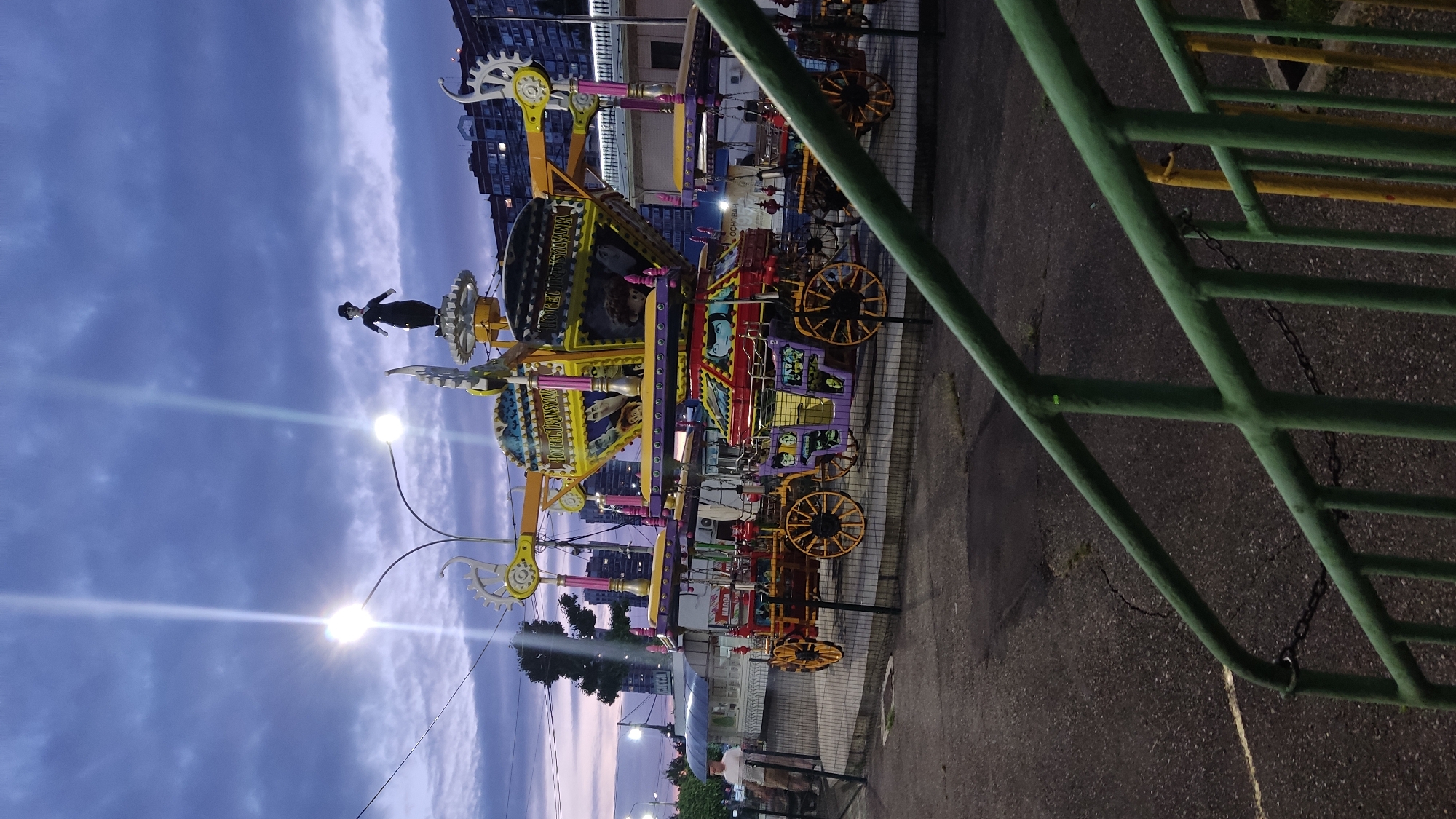 In Krasnodar, Russia, a Ferris wheel amusement ride, themed as "Hotel Transylvania," is captured at dusk. The ride features stylized wagons resembling horse-drawn carriages, adorned with characters from the animated movie. Atop the ride, a small figurine resembling Charlie Chaplin stands. The sky is a deep twilight blue with dramatic clouds and two bright streetlights casting long beams of light. The ride is enclosed by a metal fence, and in the foreground, a green railing suggests the perspective of someone observing the scene from a slightly elevated position. The background reveals a residential building, indicating an urban setting. A lone man in a light-colored shirt and dark pants stands near the fence, looking towards the amusement ride. The ground is paved with asphalt and shows signs of wear.