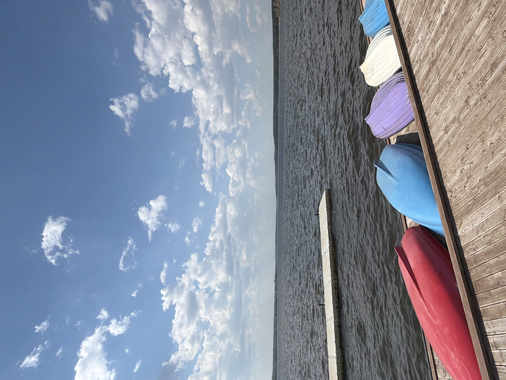 The image depicts a waterfront scene likely in the city of Votkinsk, Russia, known for its location on a large reservoir. A wooden dock extends into the water. Several colorful canoes or small boats (red, blue, lavender, white and teal) are stacked or stored along the edge of the dock. A narrow floating dock, made of light-colored material, is visible in the water. The water surface is slightly choppy. A shoreline is visible in the distance. The sky above is partly cloudy with a blue background.