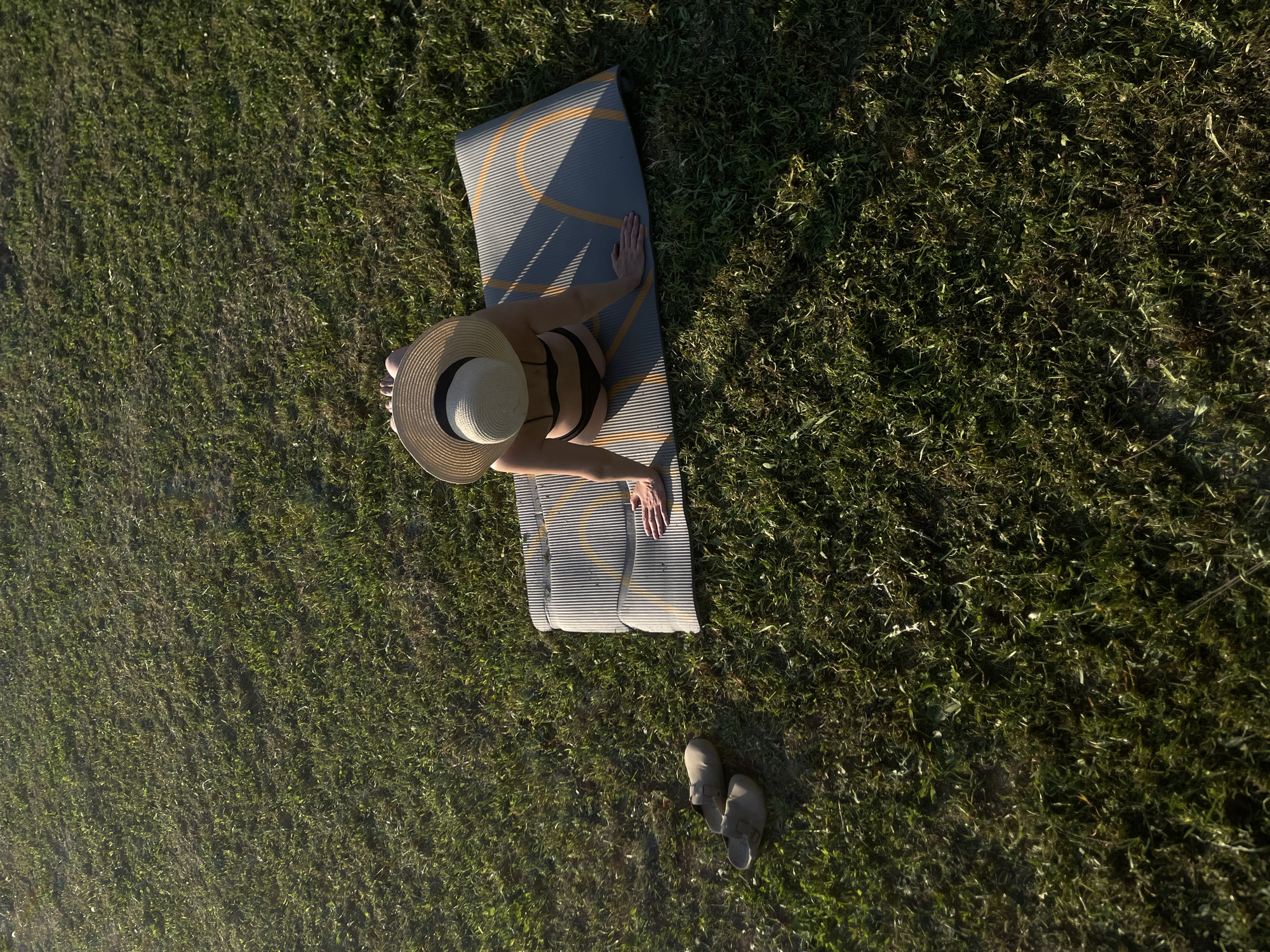A high-angle view shows a woman sitting on a gray yoga mat with yellow stripes in a grassy area in Подшивалово, Russia. She wears a black bikini and a wide-brimmed straw hat. Her bare feet are visible. A pair of gray slip-on sandals sit on the grass next to the mat. The grass is a mix of green and brownish-green hues, suggesting a natural outdoor setting.